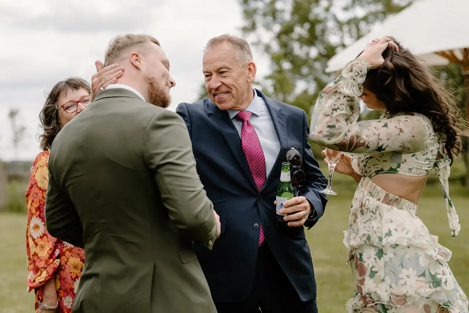 A group of five people socializing outdoors on a cloudy day. The man in the center is smiling, wearing a dark suit with a pink tie, holding a beer and sunglasses. To his left, a woman in a floral outfit with long dark hair is adjusting her hair and h
