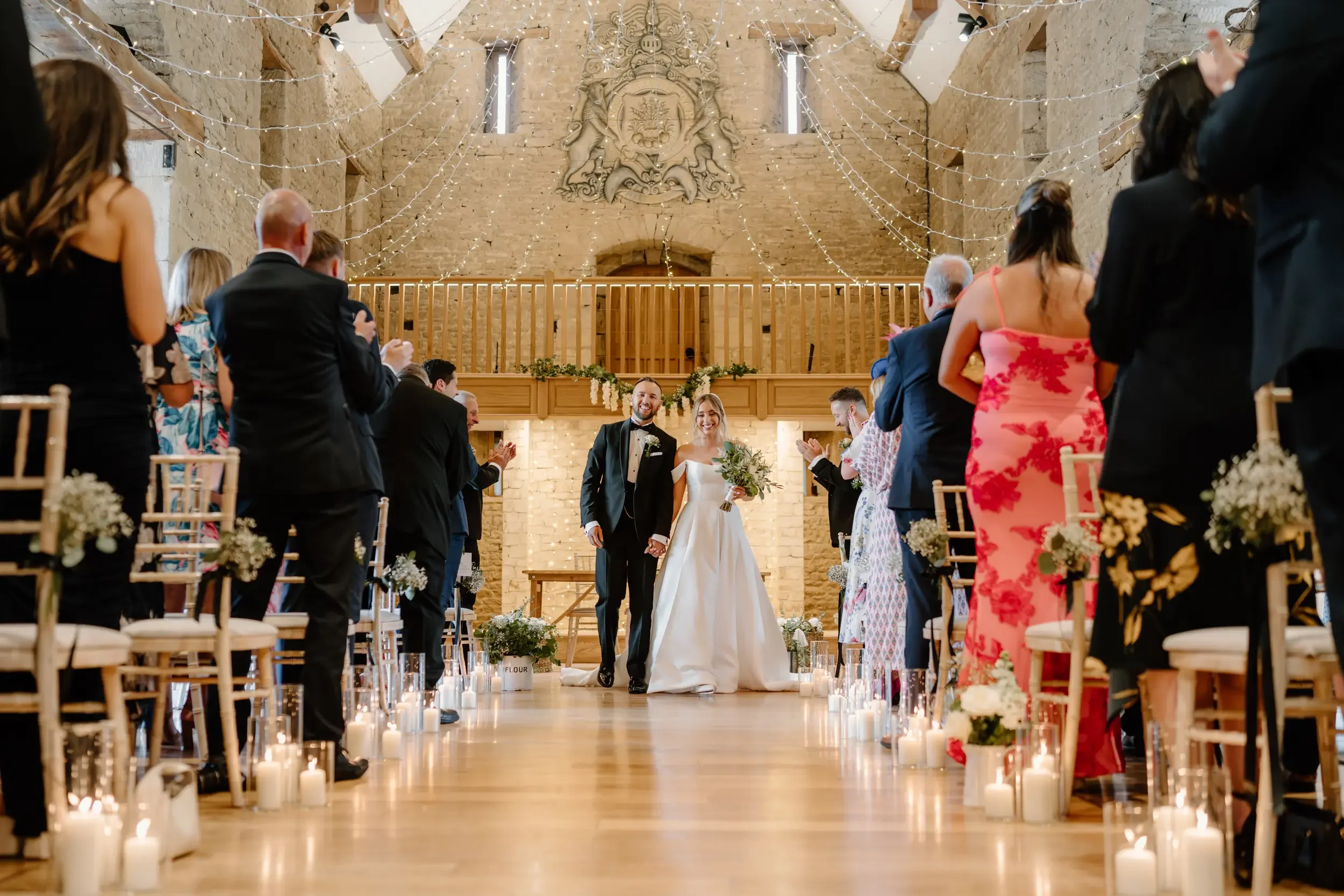 A bride and groom walk down the aisle at their wedding ceremony in a rustic venue with exposed brick walls and wooden accents, decorated with string lights and candles.