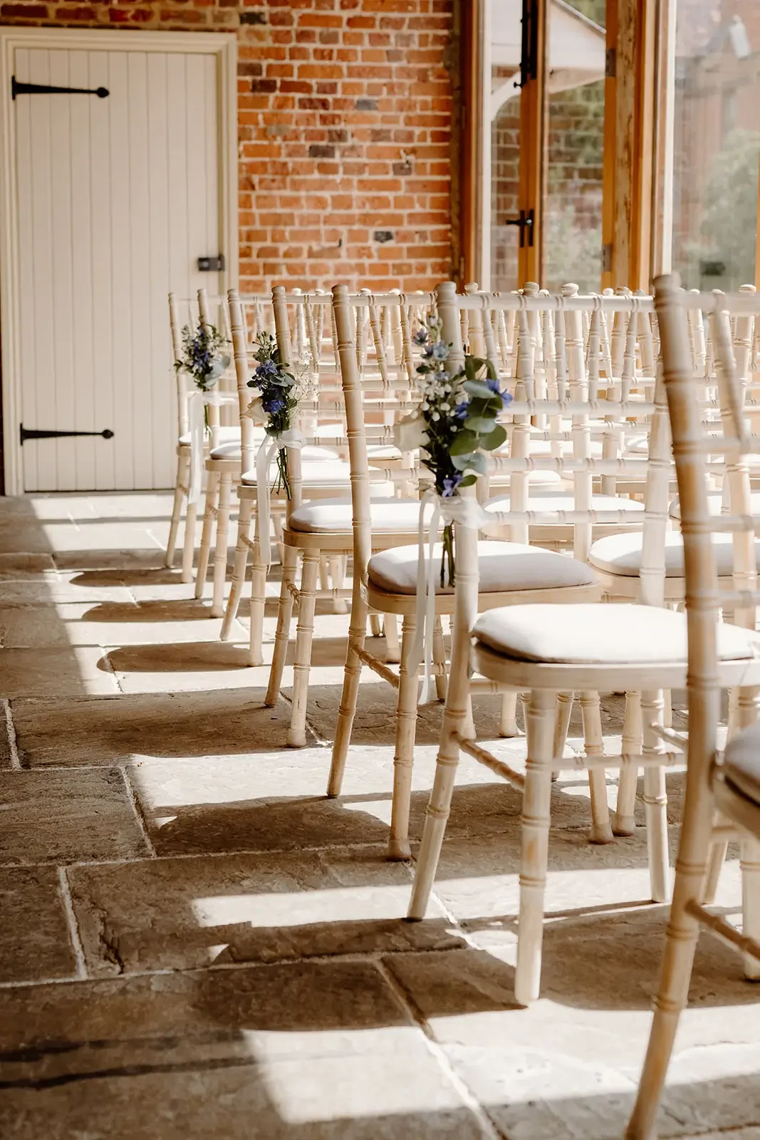 Rows of white chairs decorated with small bouquets of purple flowers and greenery, set up for a wedding or event inside a sunlit room with brick walls and large windows.