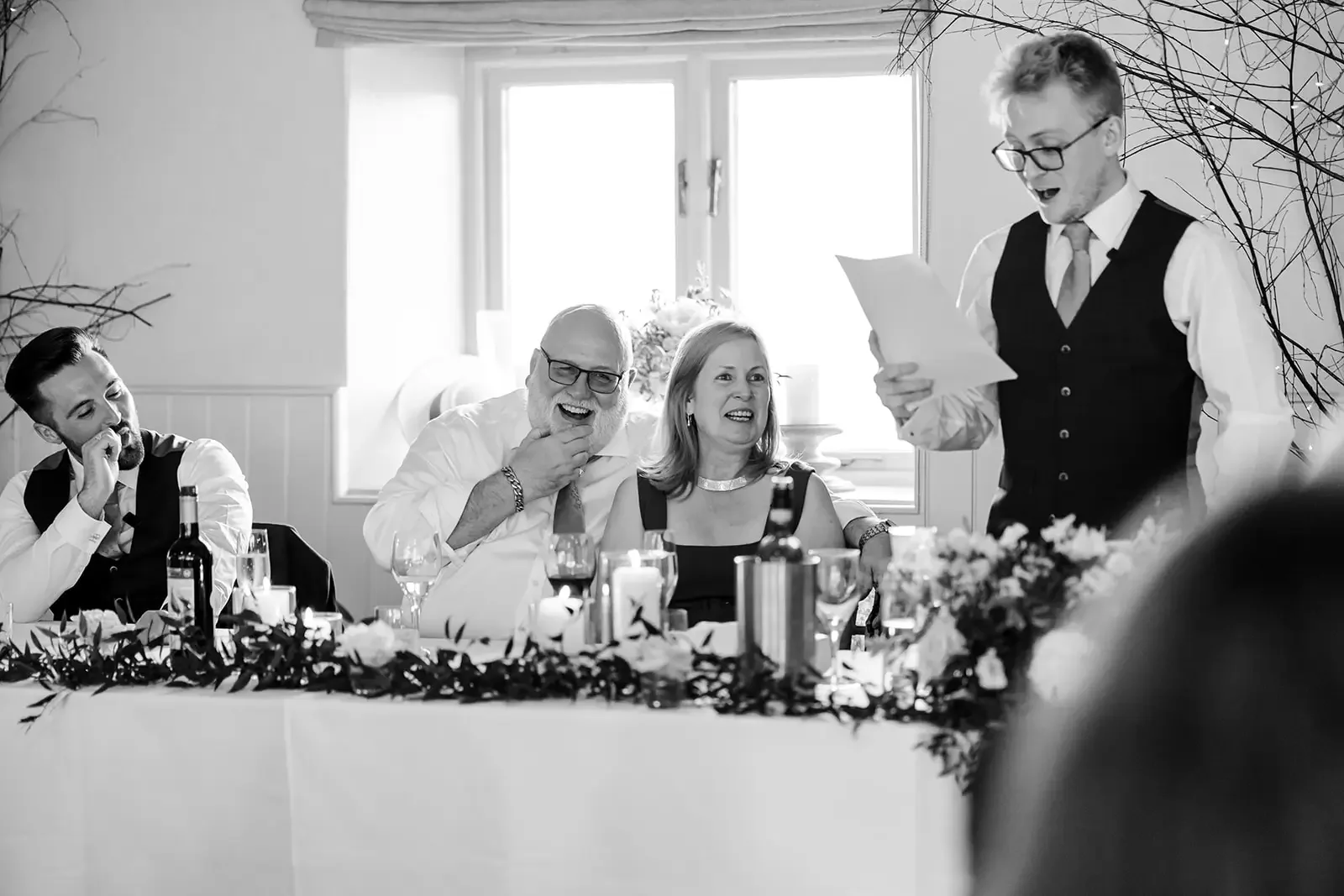 A black-and-white photo of a formal dinner event. Four people are visible, with one man standing and reading from a paper, while the others are seated at a decorated table, smiling and looking at the speaker. The table has candles, wine bottles, and 