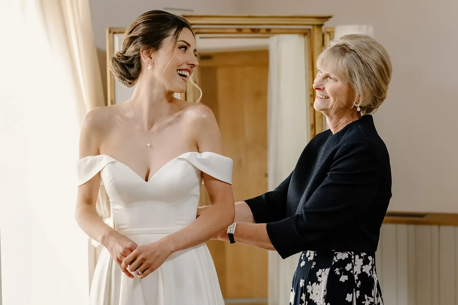 A bride in a white off-the-shoulder wedding dress smiling and holding hands with her mother, who is wearing a black blazer and floral skirt, in a warmly lit room.
