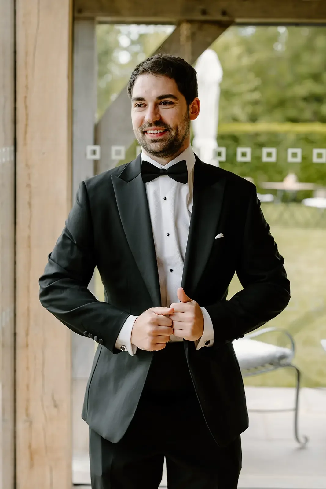 Man in a black tuxedo with a bow tie standing indoors near a glass door with a view of greenery outdoors.