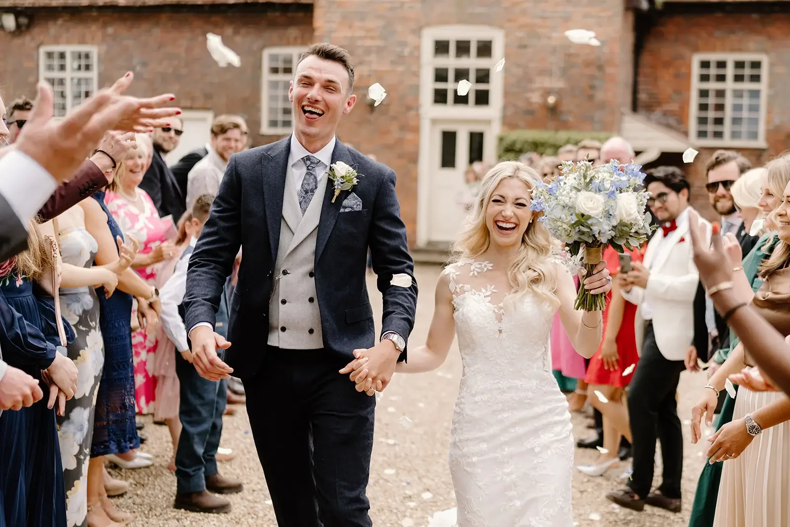 A smiling bride and groom walking hand in hand at their outdoor wedding, surrounded by wedding guests, with the bride holding a bouquet of flowers, and confetti in the air.