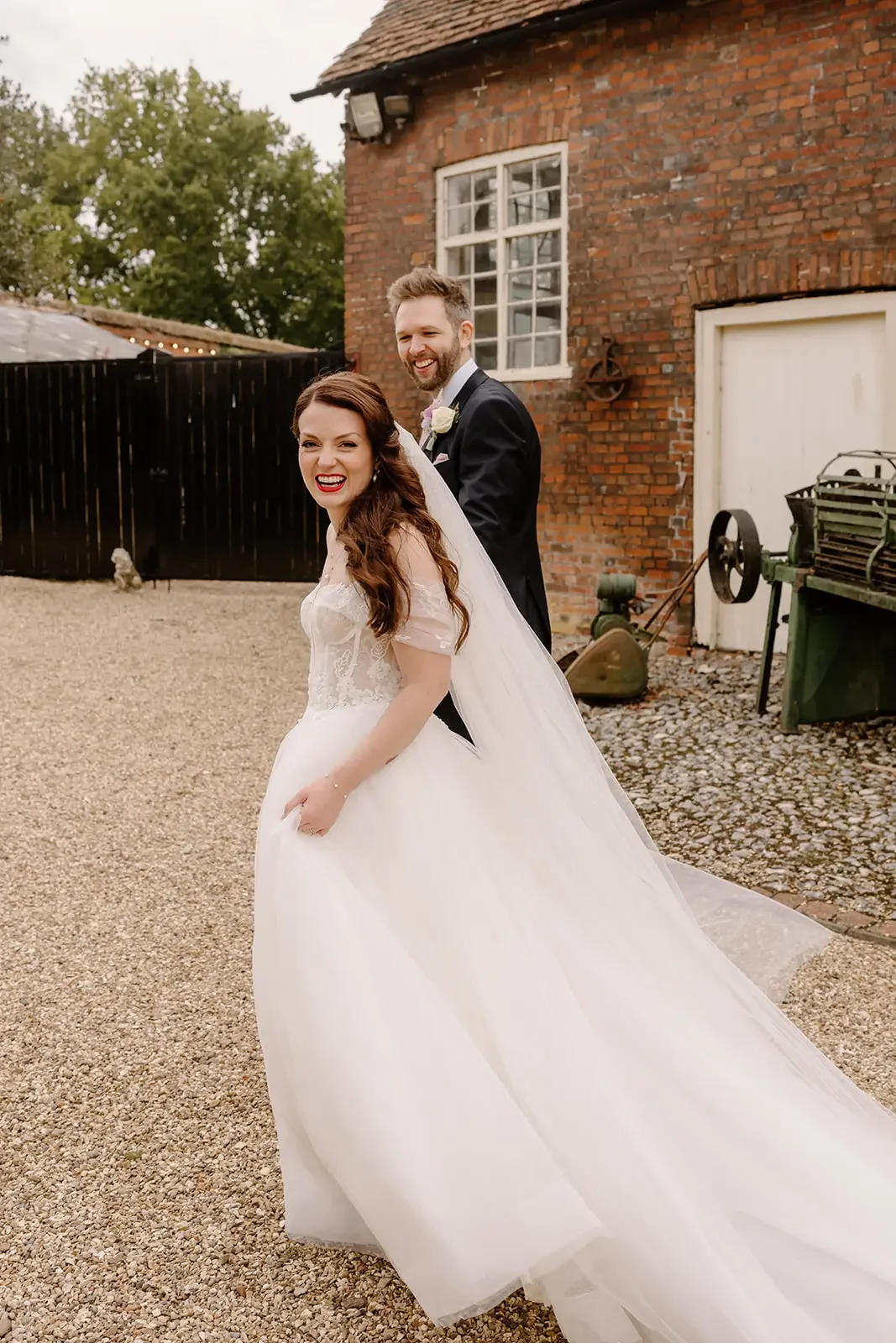 A bride and groom smiling outdoors, the bride in a white wedding dress with lace details and a long flowing train, the groom in a dark suit with a boutonniere, standing on a gravel path in front of a rustic brick building with a black fence and green