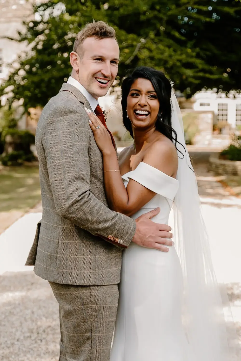 A newlywed couple smiling and laughing outdoors, with trees and buildings in the background. The man is in a brown plaid suit, and the woman is in a white off-shoulder wedding dress with a veil.