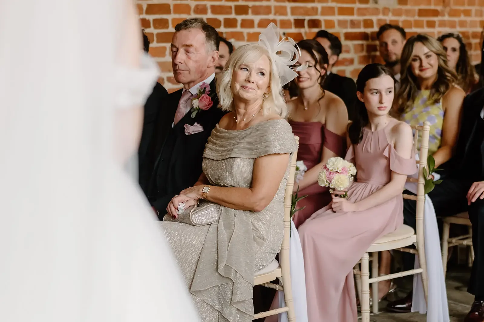 At a wedding ceremony, an older woman in a silver dress and large fascinator sits, smiling, next to an older man in a dark suit. Behind them are guests seated in chairs against a brick wall, some smiling, with a young woman in a pink dress holding a 