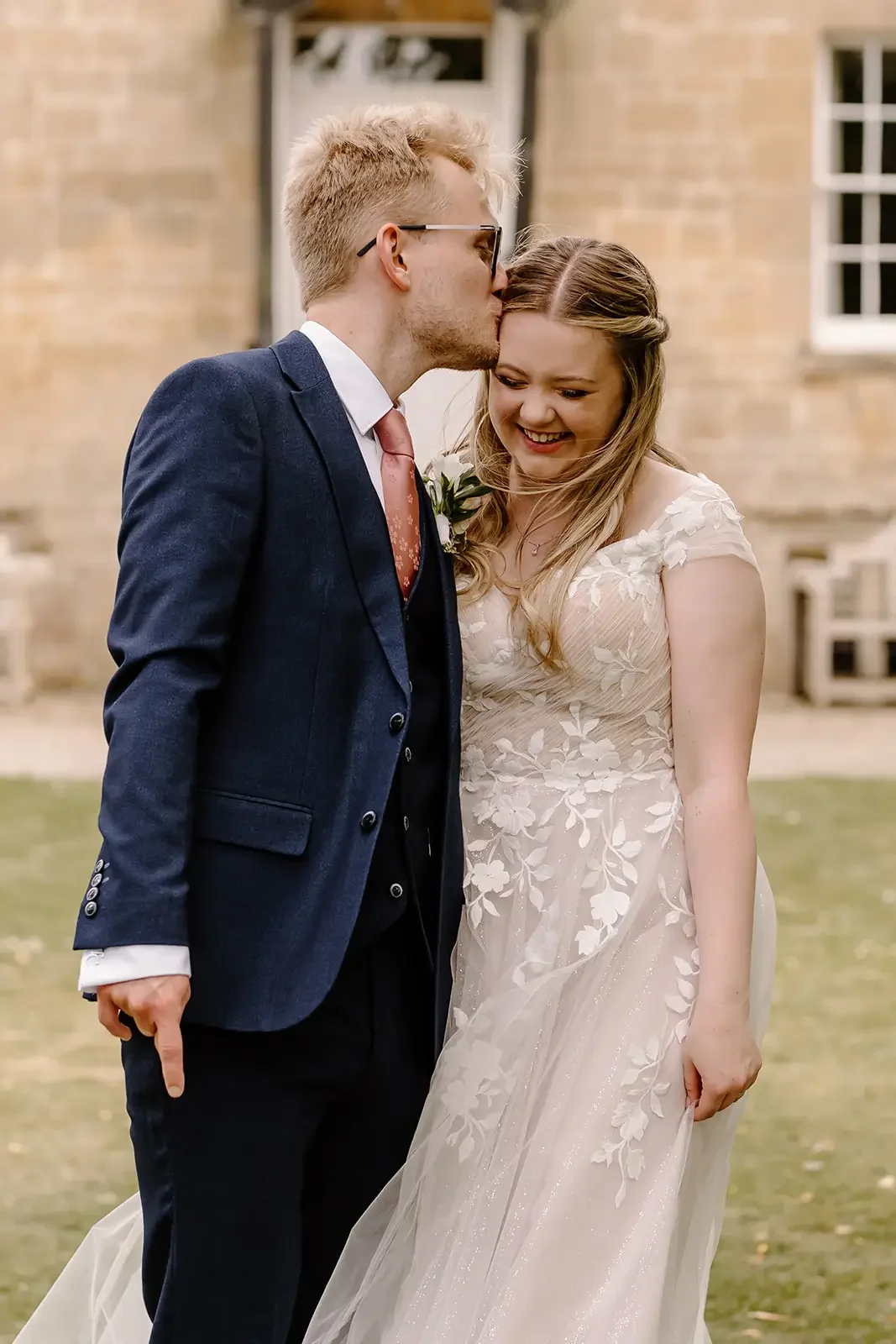 A man in a dark blue suit kissing a woman in a light-colored wedding dress on the forehead outside a brick building with windows.