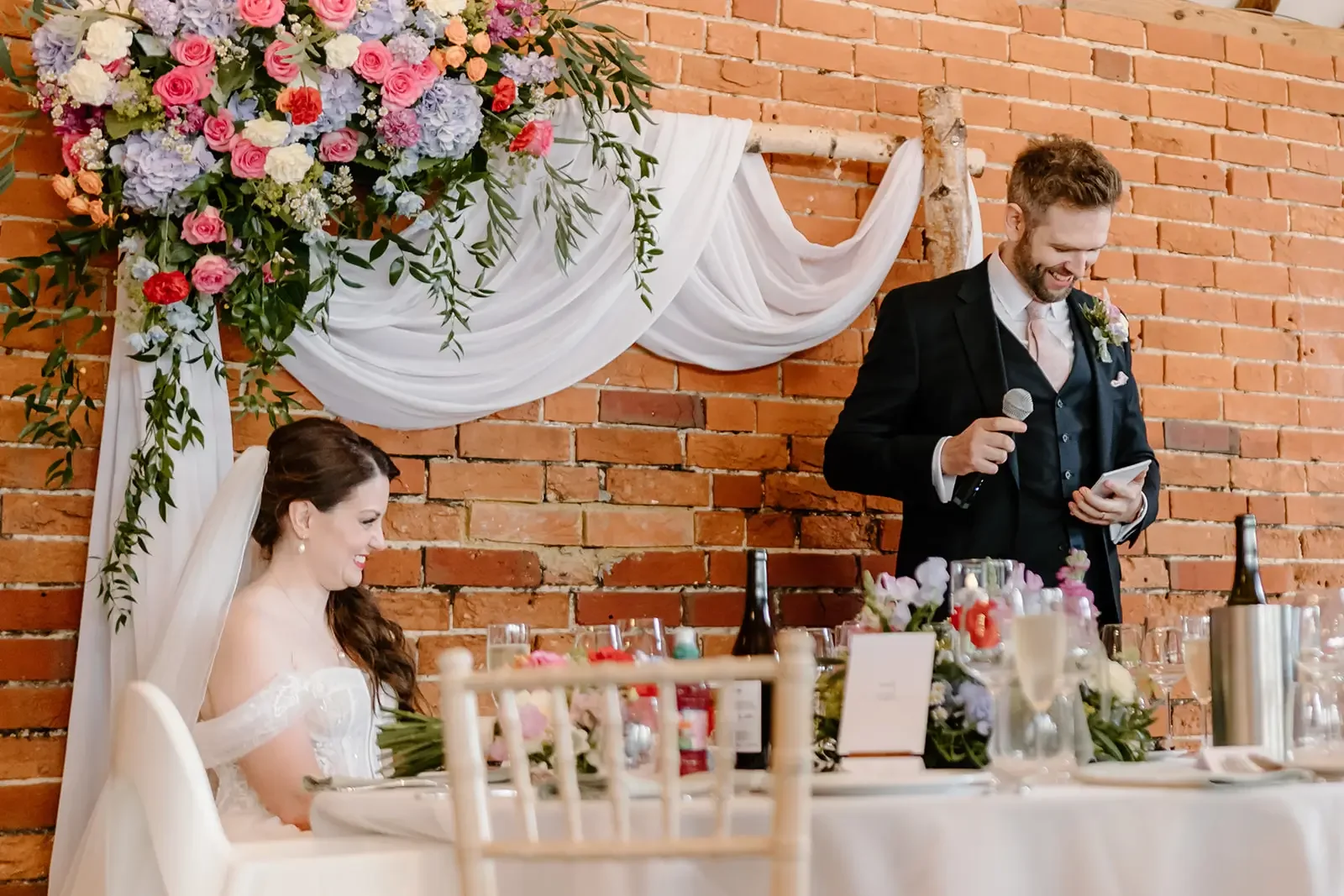 A wedding reception with a bride sitting at a table and a groom giving a toast with a microphone in front of a brick wall decorated with white drapes and pink, purple, and white flowers.