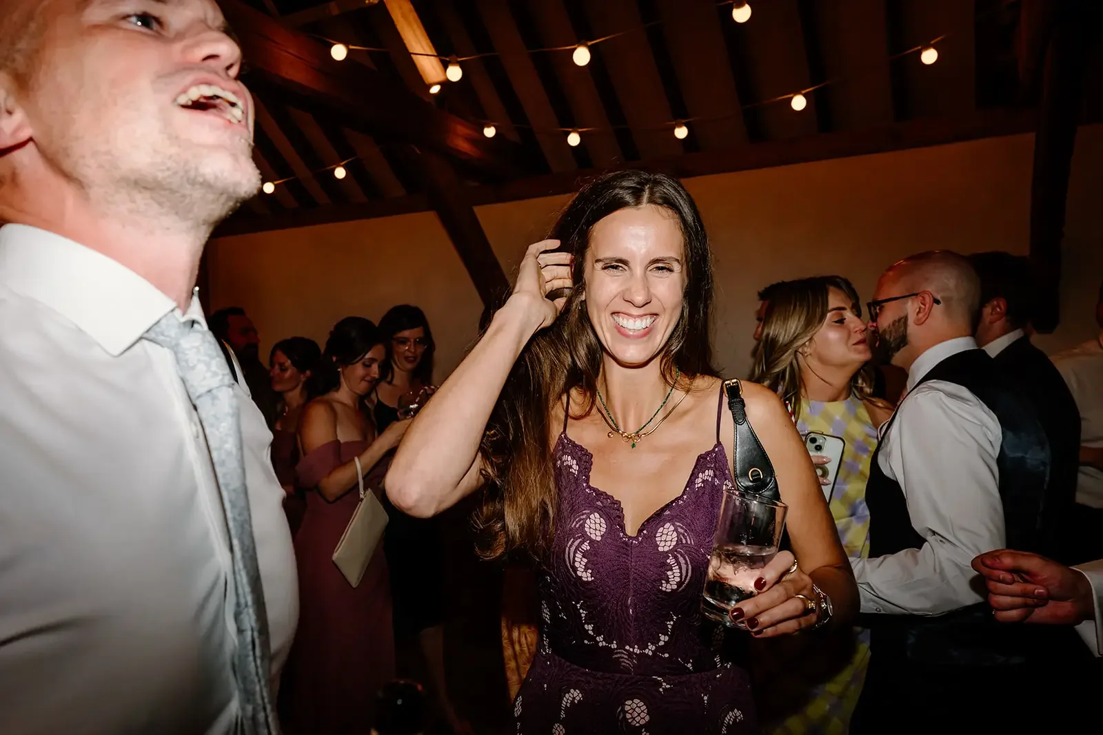 A woman with long brown hair, wearing a purple lace dress, smiling and holding a glass of water at a social gathering or party.