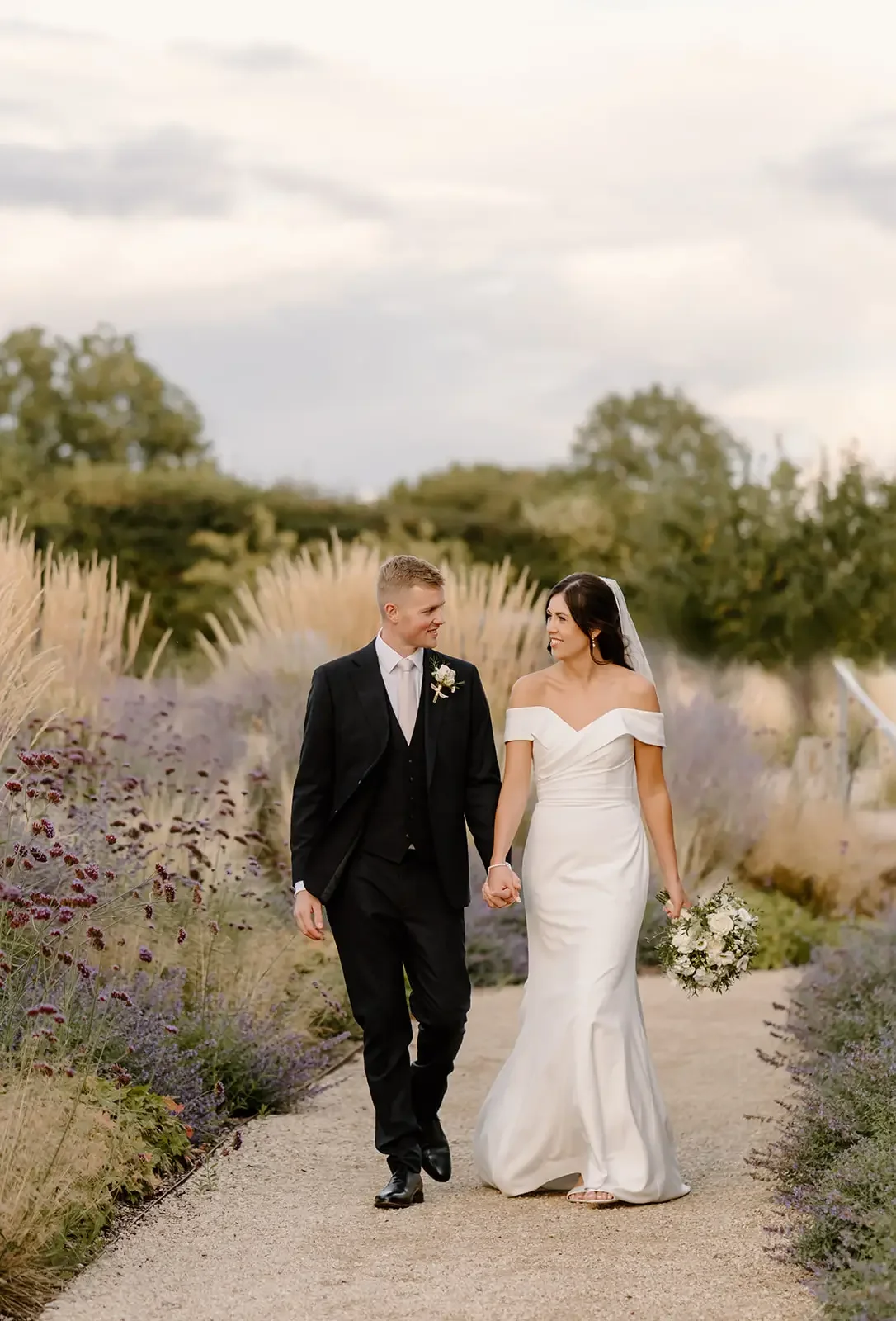 A bride and groom walking holding hands on a garden path, surrounded by purple flowers and tall ornamental grasses during the daytime.