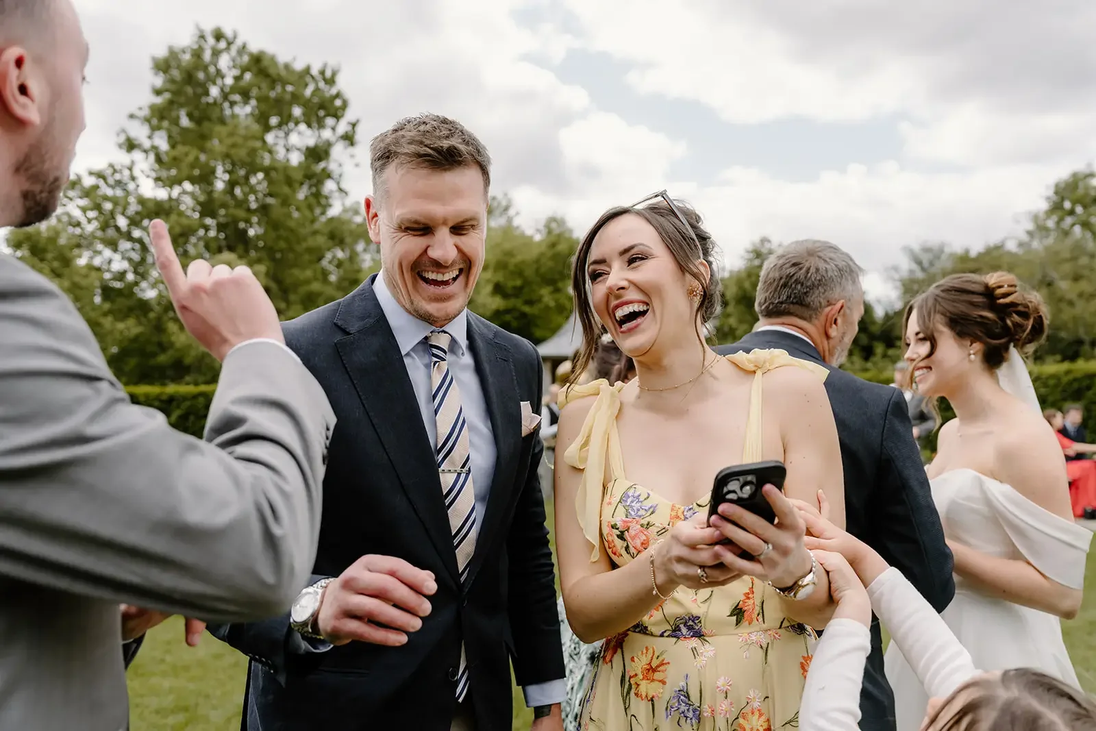 People laughing and talking outdoors at a wedding or outdoor event, with trees and cloudy sky in background.