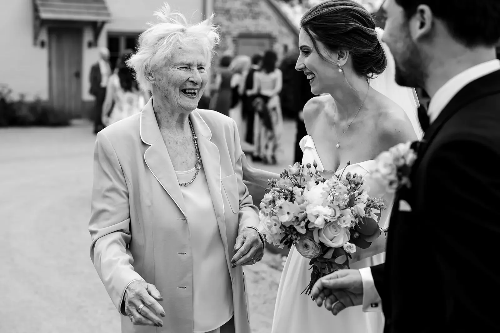 A black and white photograph of a joyful moment at a wedding, featuring an elderly woman smiling and talking with a bride who is holding a bouquet of flowers, and a groom partially visible on the right. In the background, guests are socializing outdo