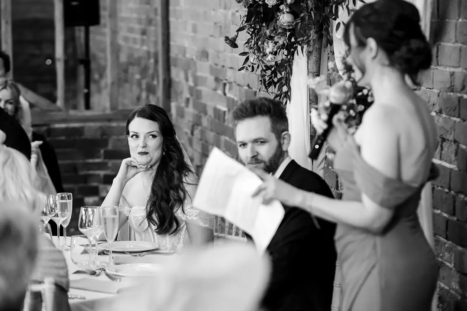 A wedding reception with a woman in bridal attire listening to a woman giving a speech, while a man with a beard listens, seated at a decorated table.