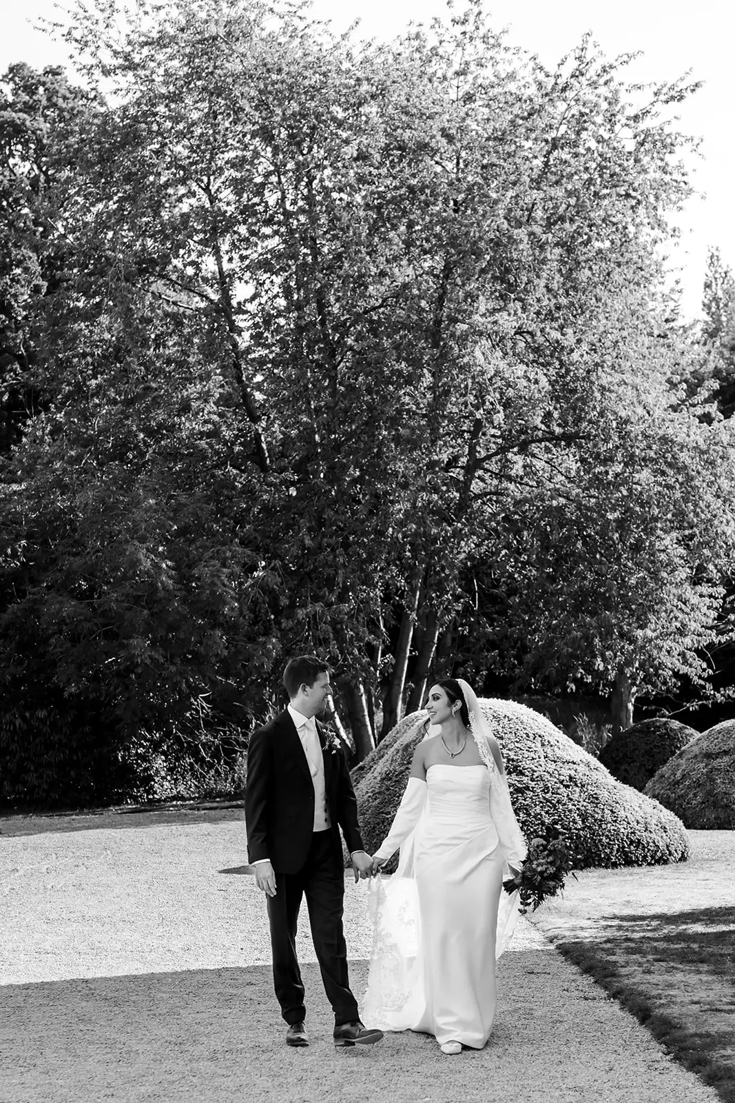 A black and white photograph of a bride and groom walking hand in hand outdoors, with trees and bushes in the background.