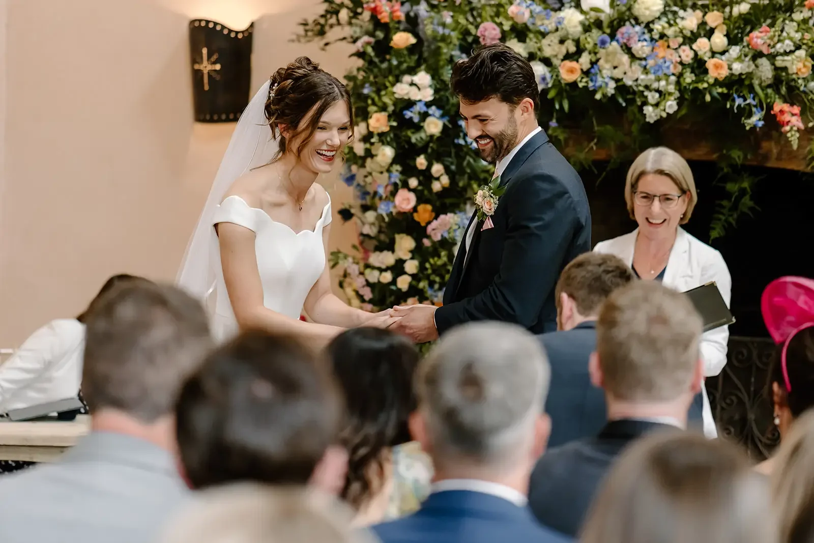 Bride and groom exchanging vows during a wedding ceremony, with a floral backdrop and guests watching.