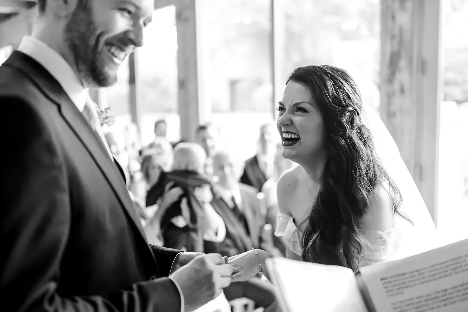 A bride and groom exchanging rings during their wedding ceremony, smiling and laughing inside a bright, airy venue.