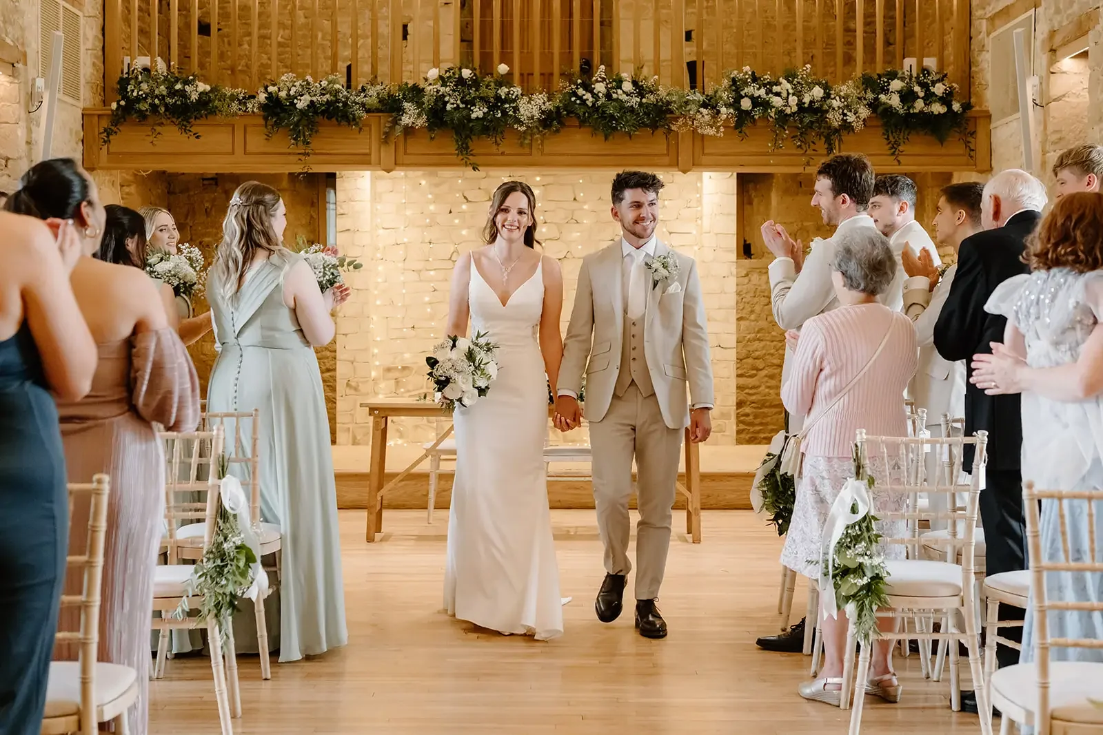 A wedding ceremony with a bride and groom holding hands, walking down the aisle in a decorated venue, surrounded by seated guests clapping and smiling.