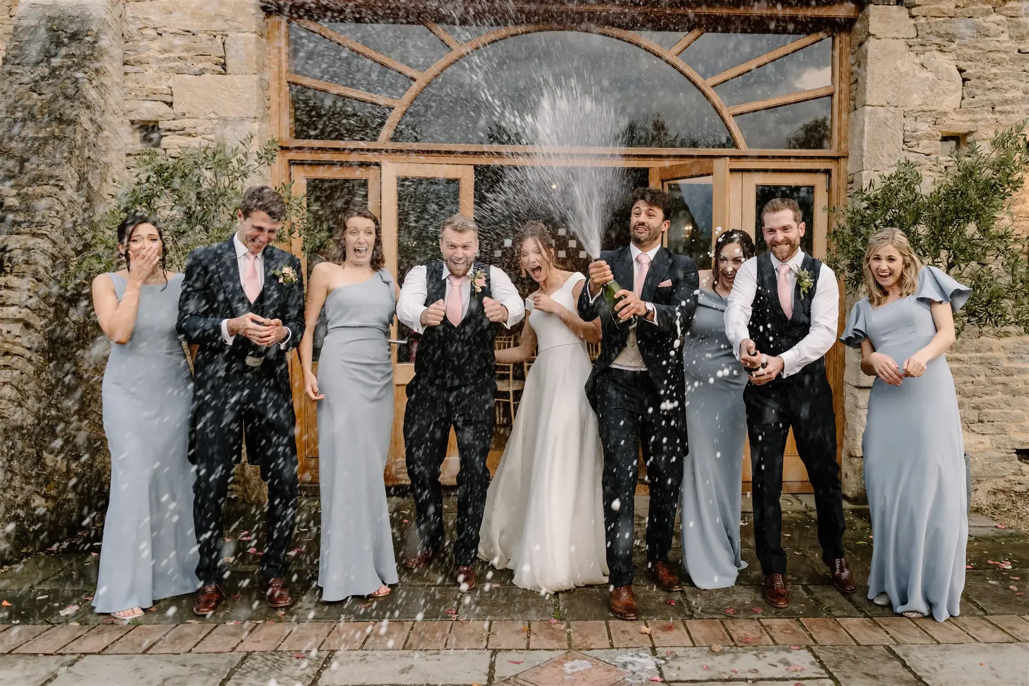 A wedding celebration with nine people, including a bride and groom, standing outside a stone building with a large wooden door and glass windows. They are cheering and spraying champagne, with joyful expressions and formal attire, including dresses 