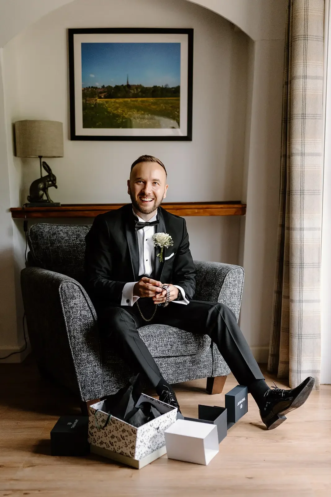 A man in a tuxedo sitting in a gray armchair in a room with a framed landscape picture on the wall behind him. He is smiling, holding a necklace, and has a white boutonnière on his tuxedo. There are gift boxes on the floor in front of him.