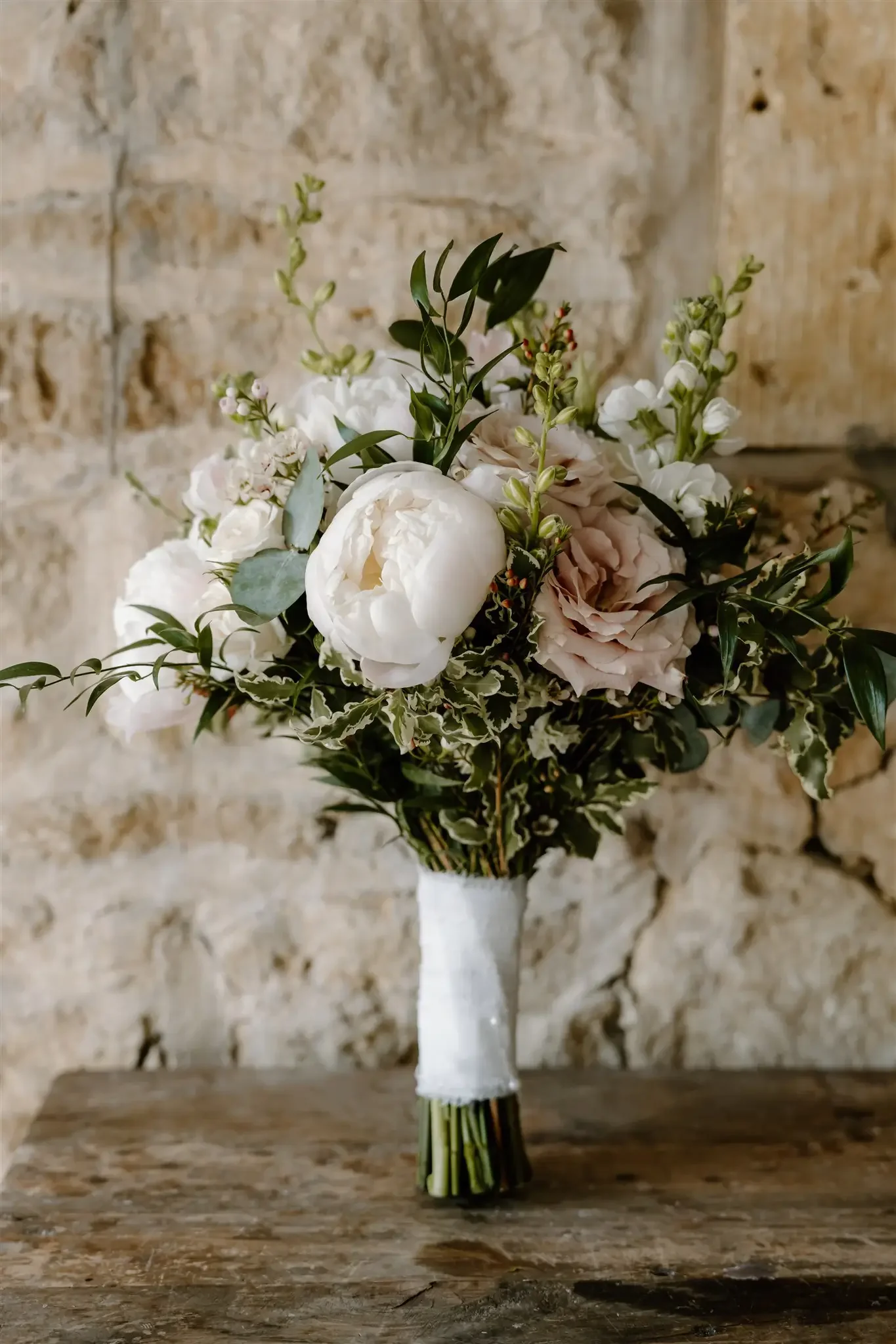 A floral bouquet in a white wrapped vase on a rustic wooden table against a stone wall background.
