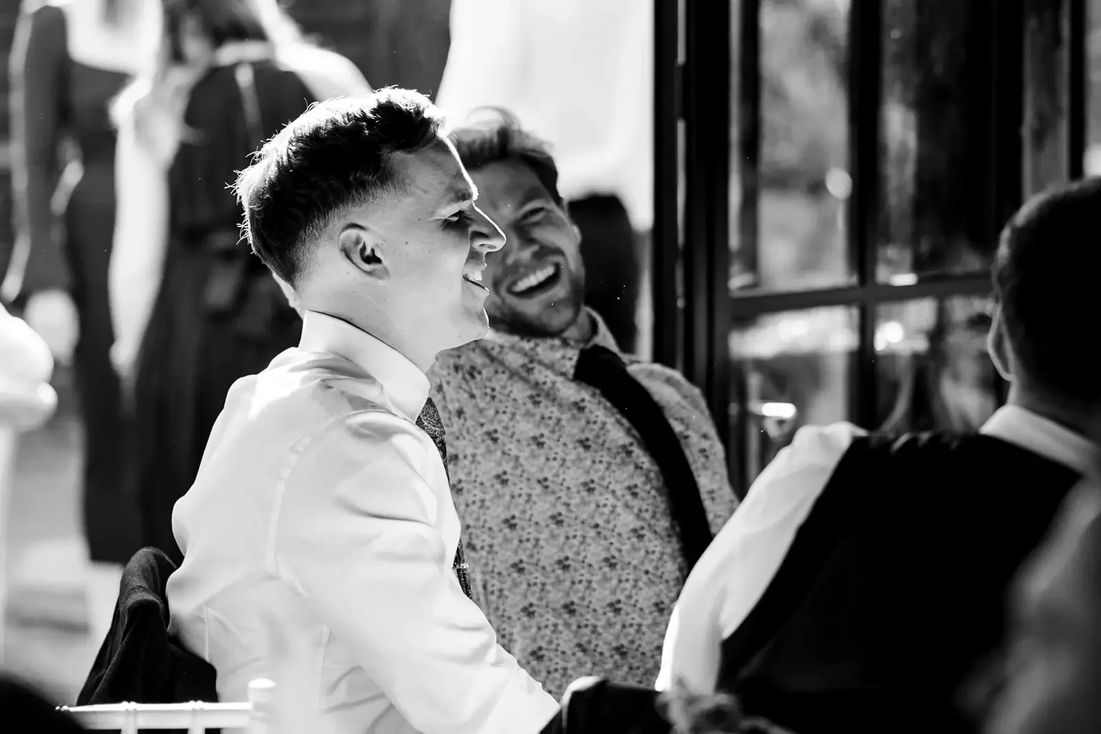 Black and white photo of three young men in formal attire smiling and laughing together indoors near a window.