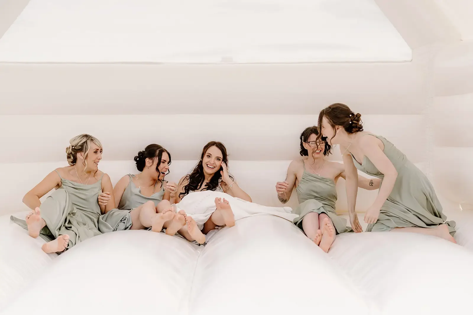 Six women in matching light green dresses sitting and laughing on a white inflatable bed.