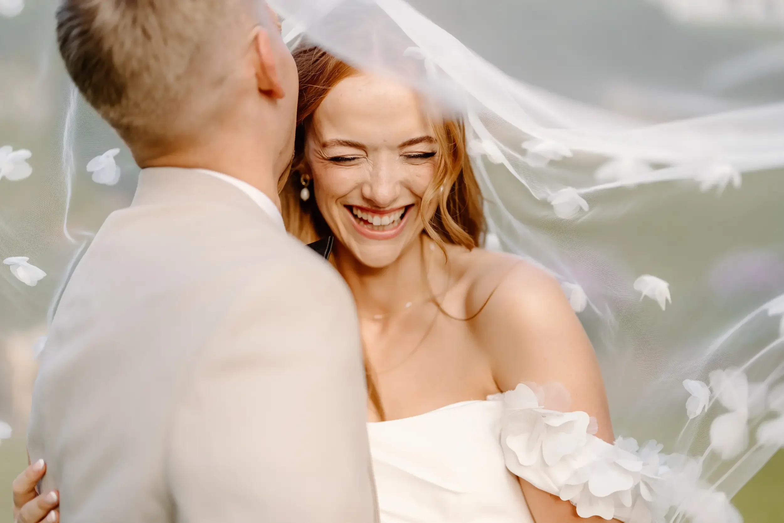 A smiling red-haired bride in a wedding dress with a veil, and a groom in a beige suit, embracing outdoors.