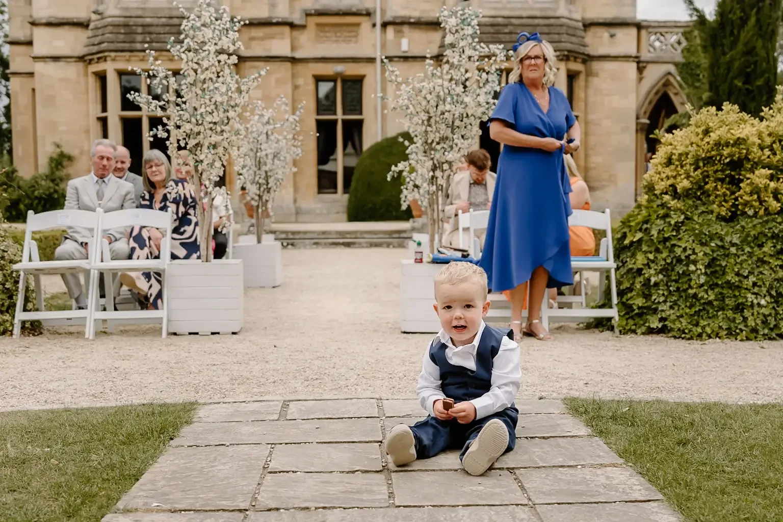 Young boy in formal attire sitting on stone pathway at outdoor wedding ceremony, with guests seated on white chairs and trees decorated with white flowers in the background.