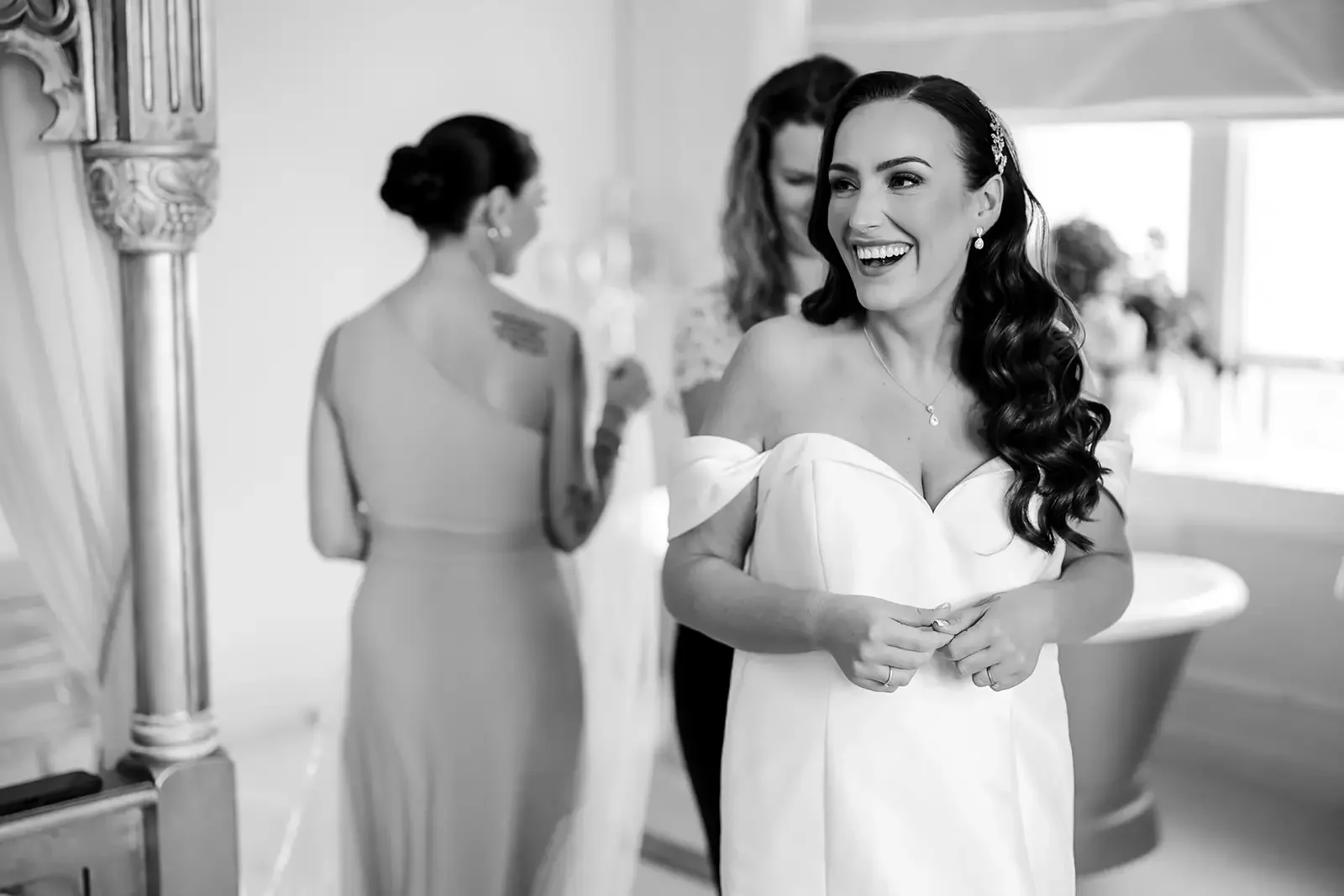 A smiling bride in a white off-shoulder wedding dress with long dark hair, standing indoors with two women in bridesmaid dresses in the background.