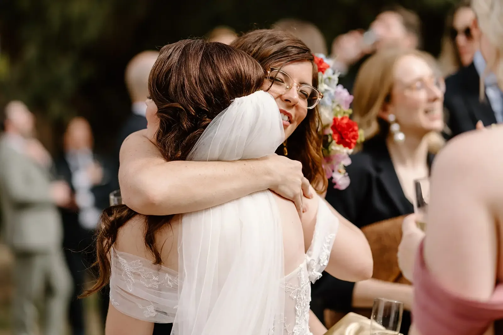 Two women hugging at a wedding reception, one in a wedding dress and glasses, the other in an off-the-shoulder dress, surrounded by guests with flowers and glasses.