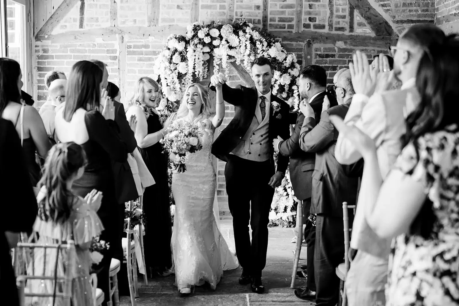 A black and white photo of a newlywed couple walking down the aisle at their wedding celebration. The bride is in a wedding dress holding a bouquet and smiling, while the groom is in a suit lifting her hand in celebration. Guests are on either side c
