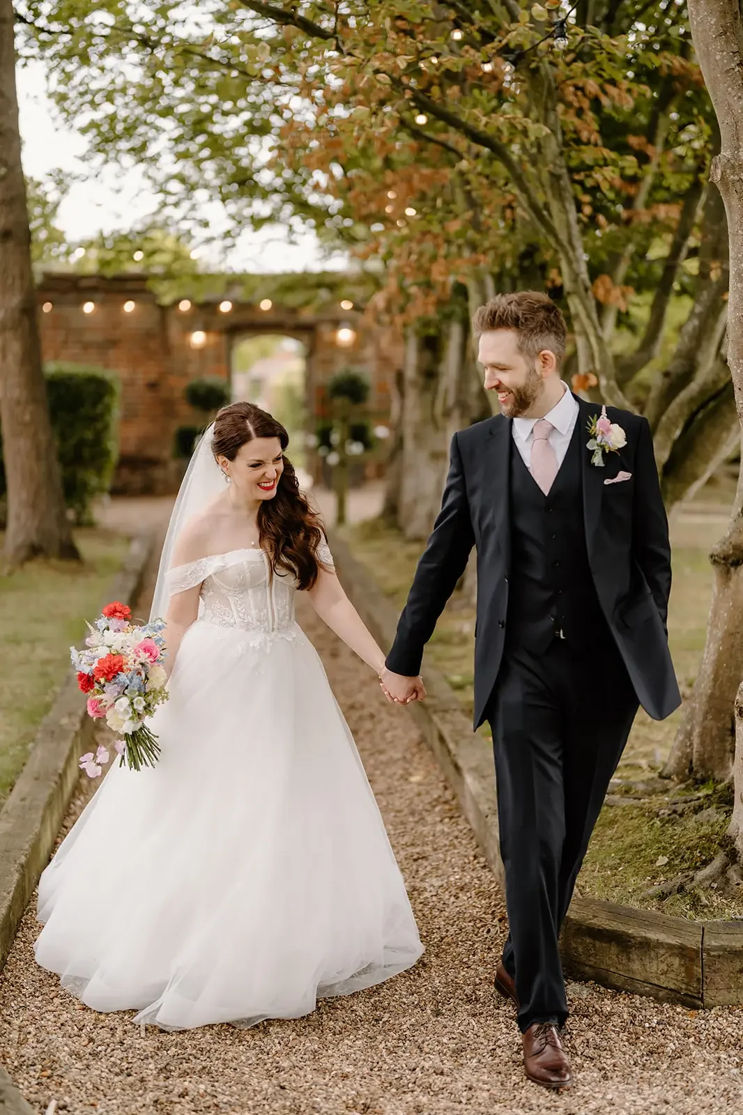 A bride and groom holding hands and walking outdoors on a gravel path, smiling at each other. The bride is wearing a white wedding dress and holding a colorful bouquet. The groom is dressed in a black suit with a light pink tie and boutonniere. There