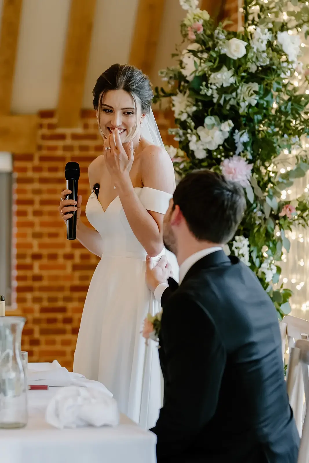 A bride in a white wedding dress holding a microphone, smiling with her hand near her mouth, while a groom in a black tuxedo kneels in front of her, holding her arm during a wedding speech or vow exchange, surrounded by flowers and warm lighting.