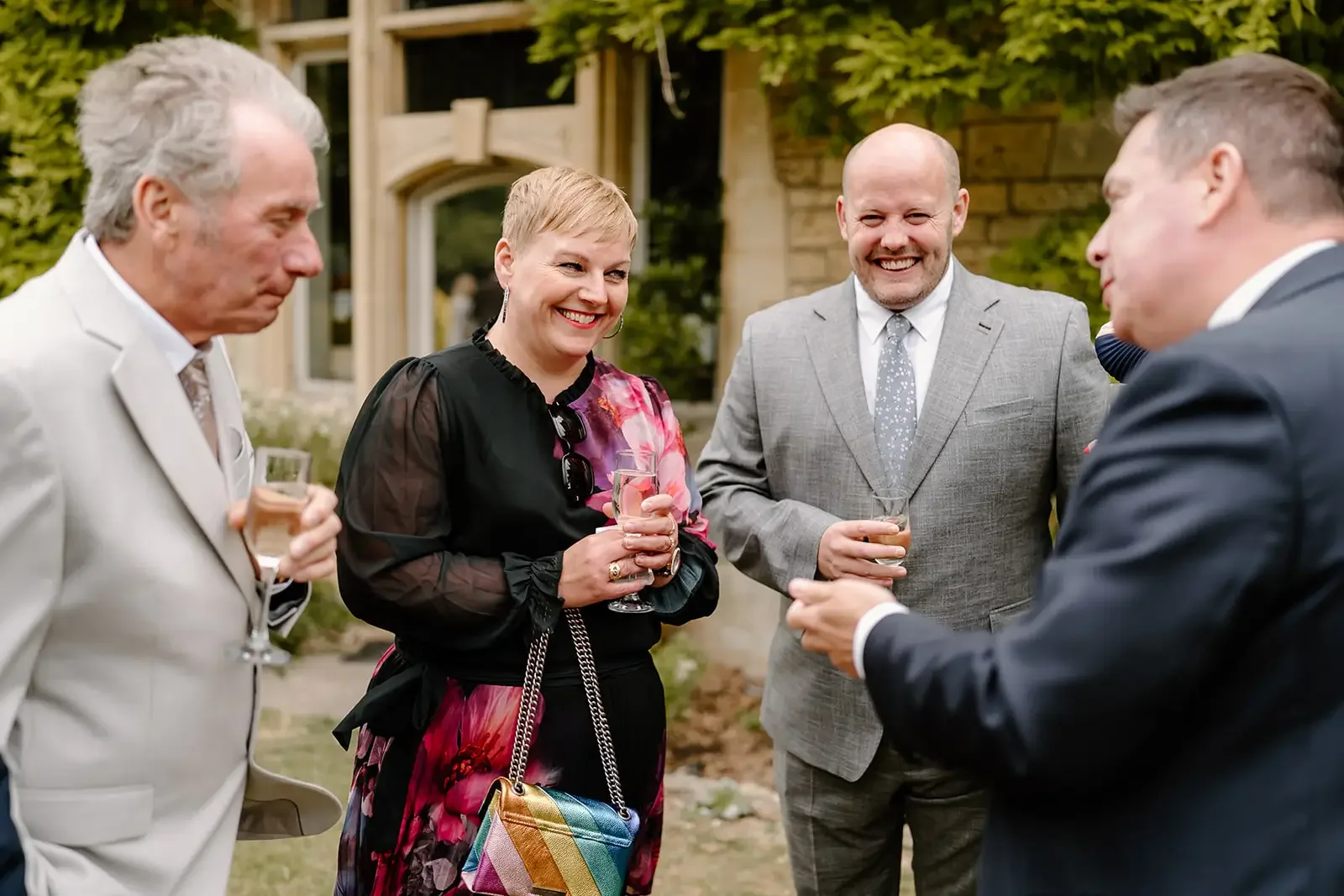 Group of five adults at an outdoor gathering, holding drinks and having a conversation, with a garden and house in the background.
