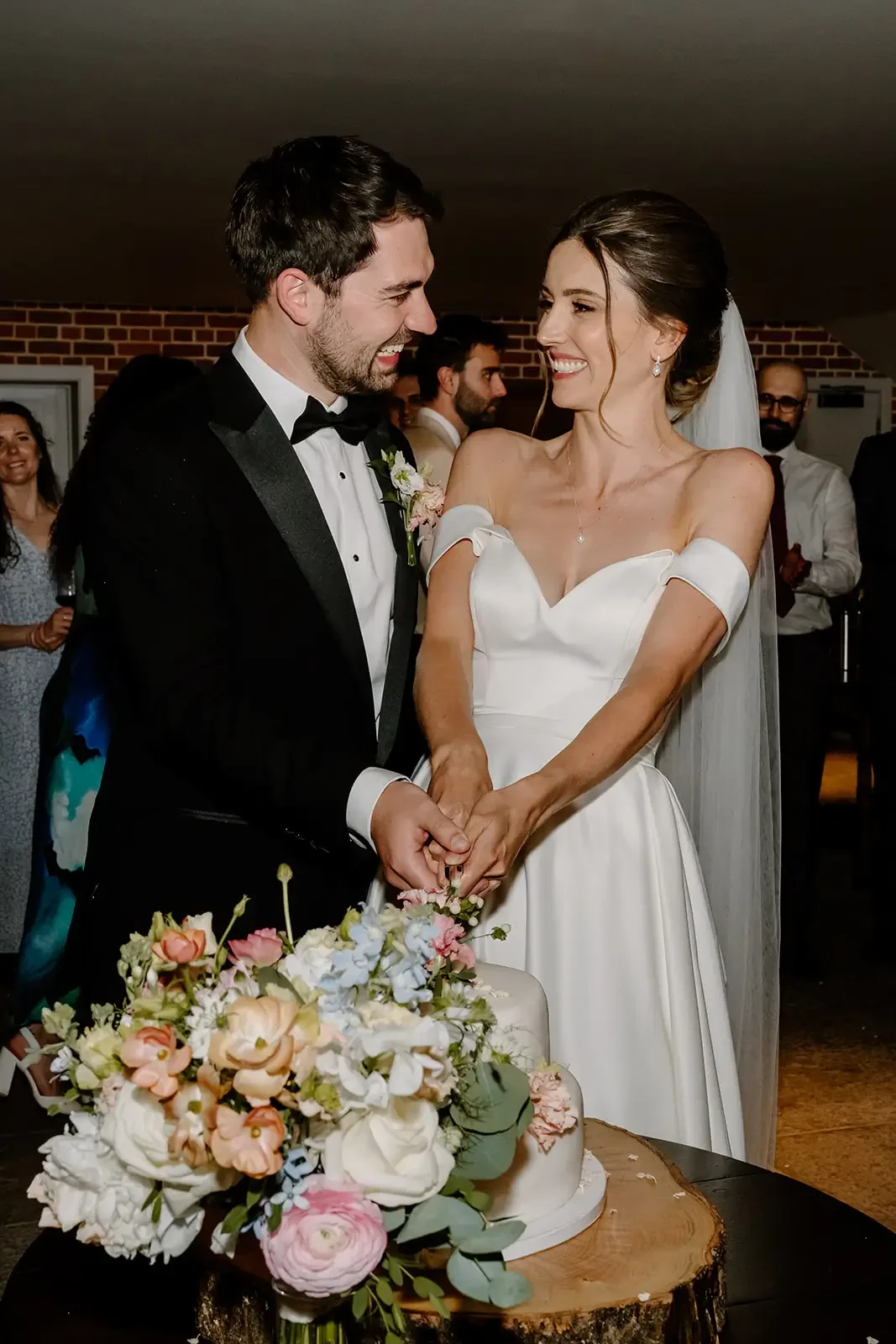 Bride and groom smiling while cutting wedding cake, surrounded by guests.