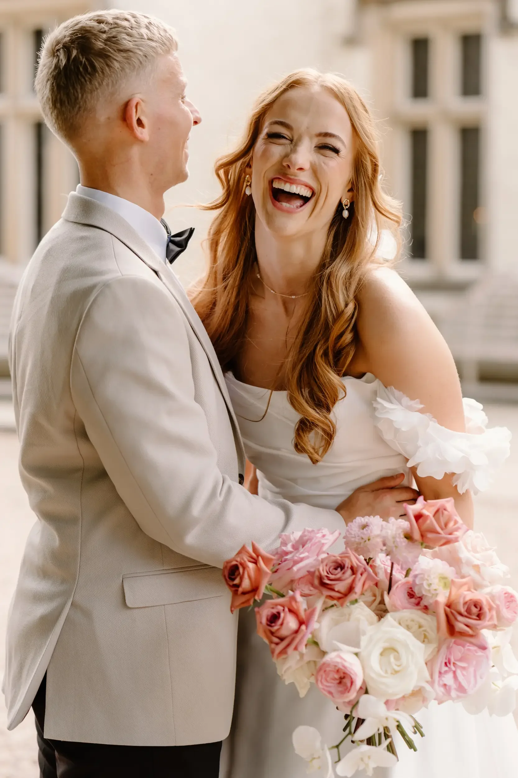 A bride and groom laughing together on their wedding day. The bride is holding a bouquet of pink and white roses, and they are standing outside a historic building.