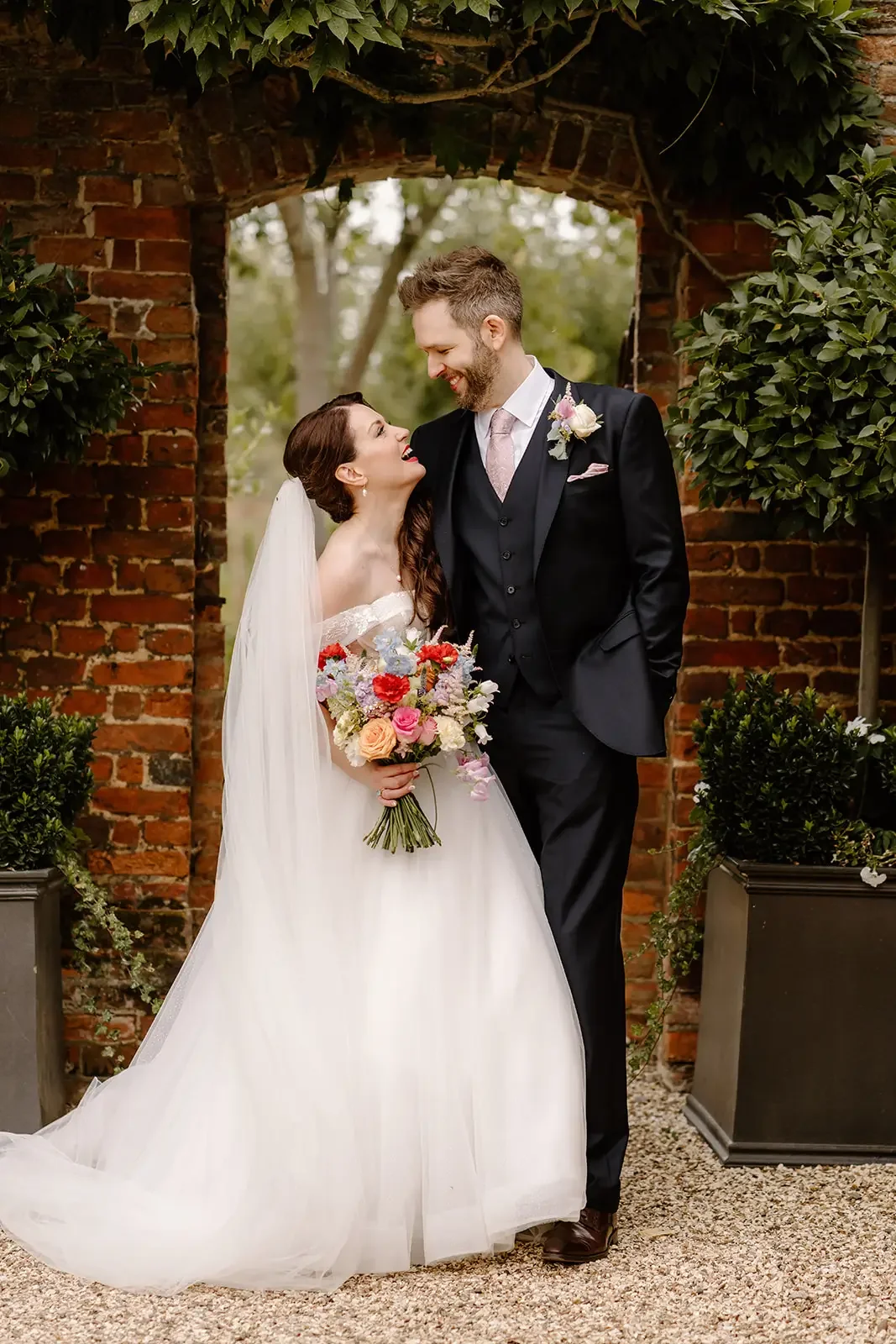 A bride and groom share a joyful moment outdoors under a brick archway surrounded by greenery. The bride is in a white wedding gown holding a colorful bouquet, and the groom is in a black suit with a pink tie and boutonniere.