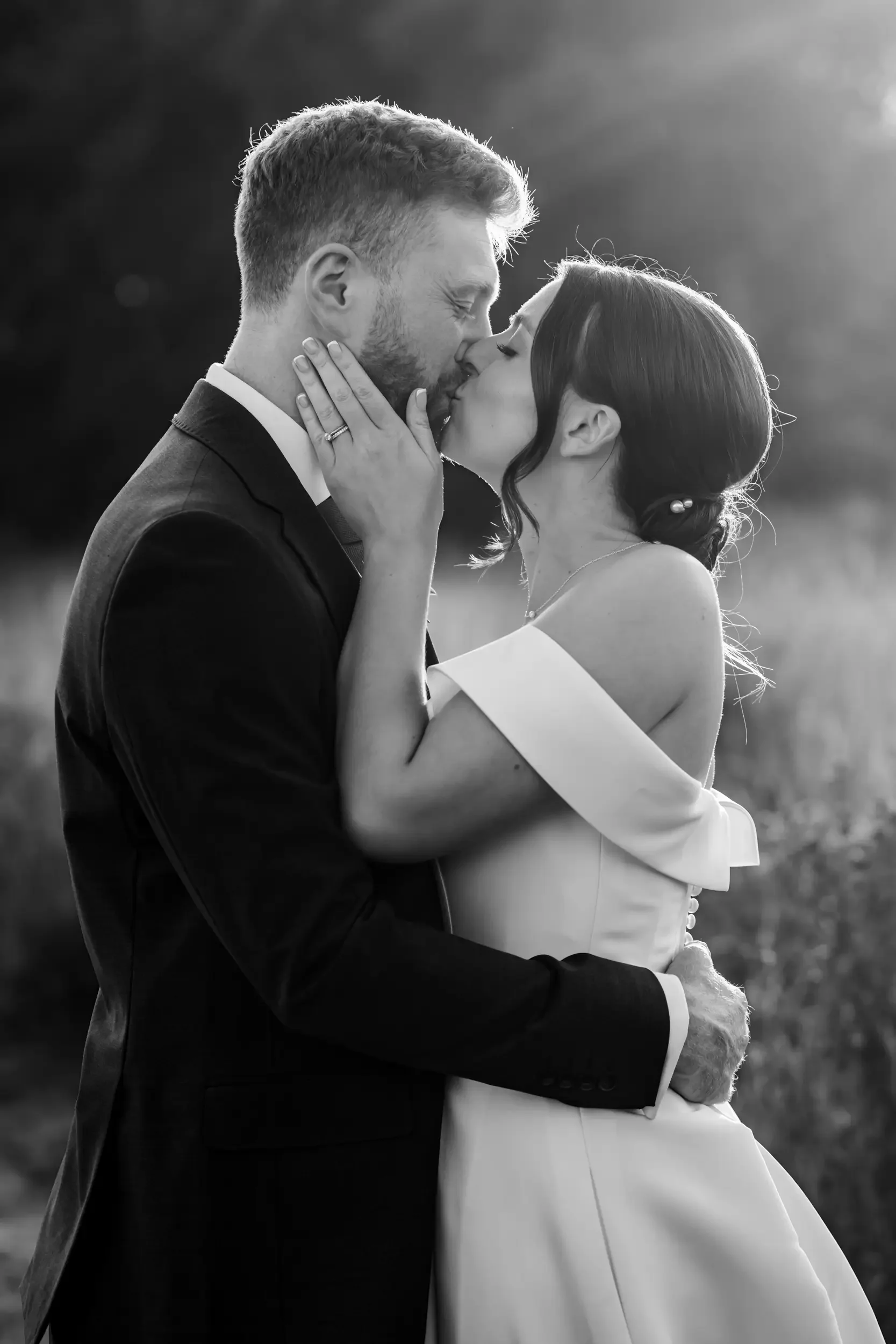 A black and white photo of a bride and groom kissing outdoors, with the bride's hand on the groom's face and his hand on her waist, during sunset.