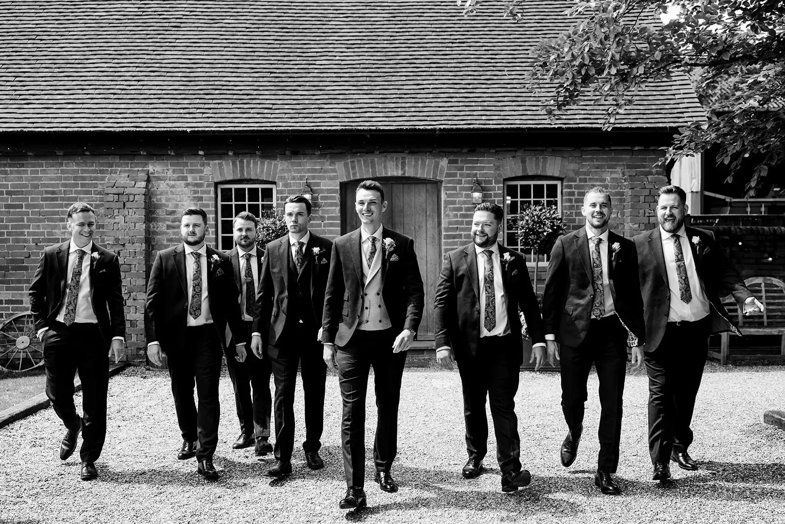 Groom and groomsmen walking outdoors in formal suits, smiling, in front of a brick building with windows and a wooden door, with trees and a bench nearby, in black and white.