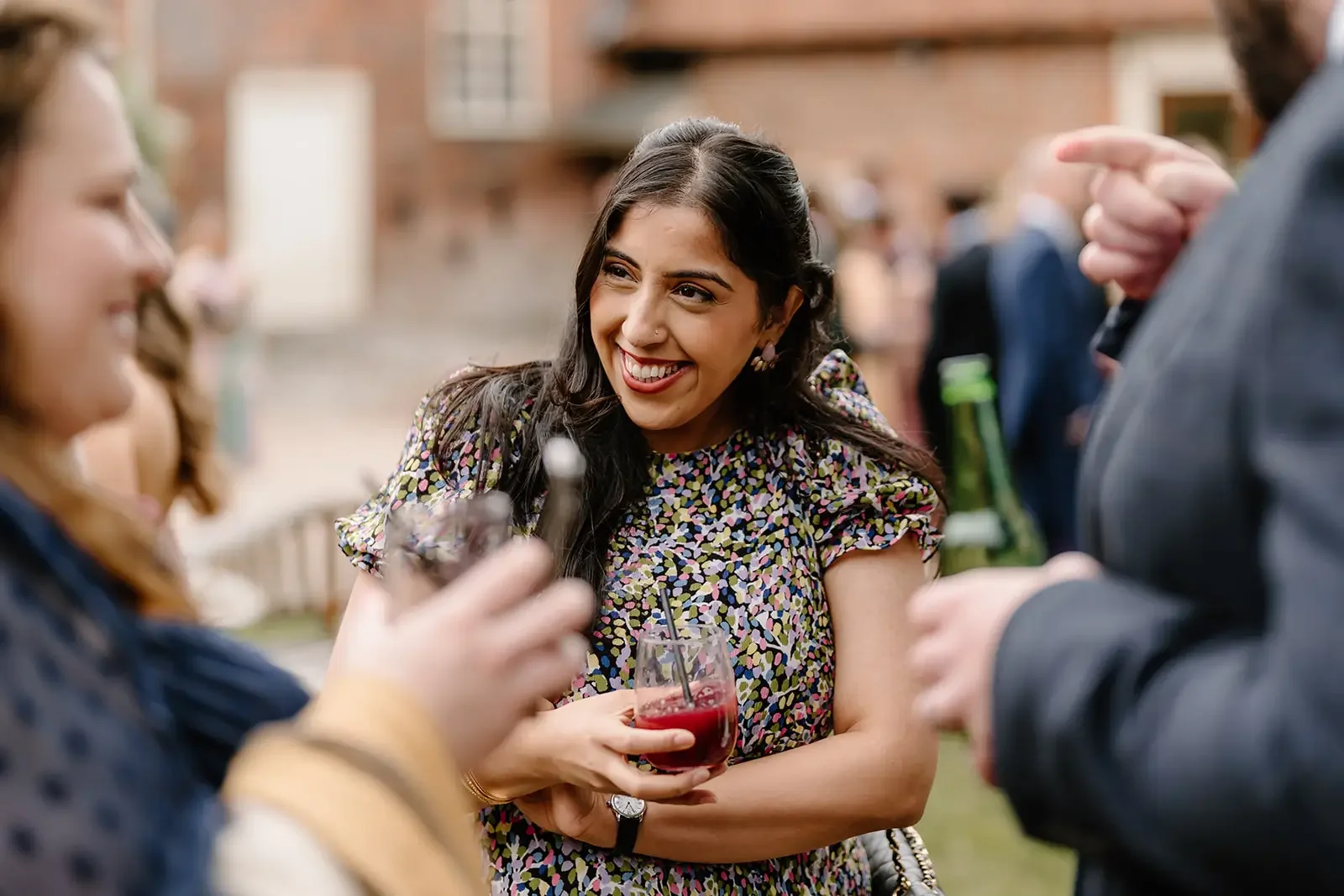 A woman with long dark hair smiling and holding a drink at an outdoor social gathering, surrounded by other people.