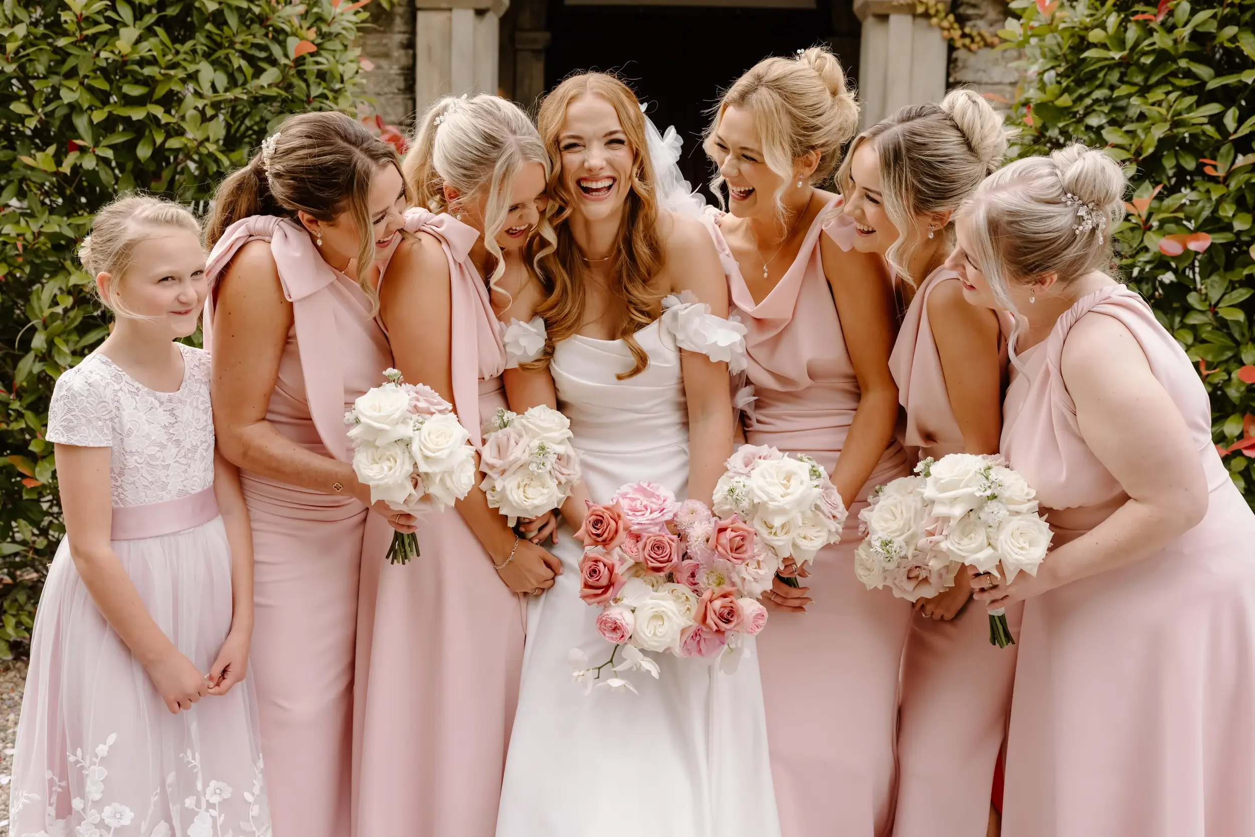 A group of women and one young girl in pink dresses standing outdoors. The women are holding bouquets of white and pink flowers and are smiling and laughing, with the bride in a white dress at the center. Green bushes are in the background.