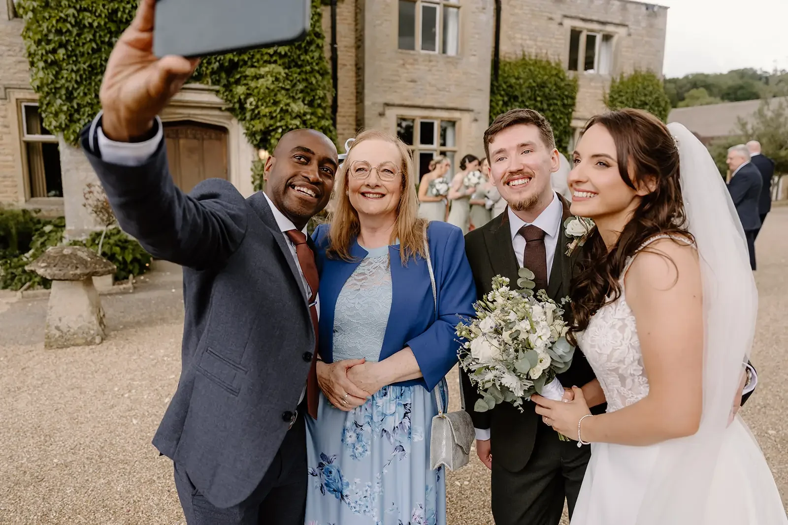 Group of five people taking a selfie outside a stone building during a wedding. The bride is holding a bouquet of white flowers and wearing a white wedding dress. The groom and another man are dressed in suits. An elderly woman in a blue outfit is sm
