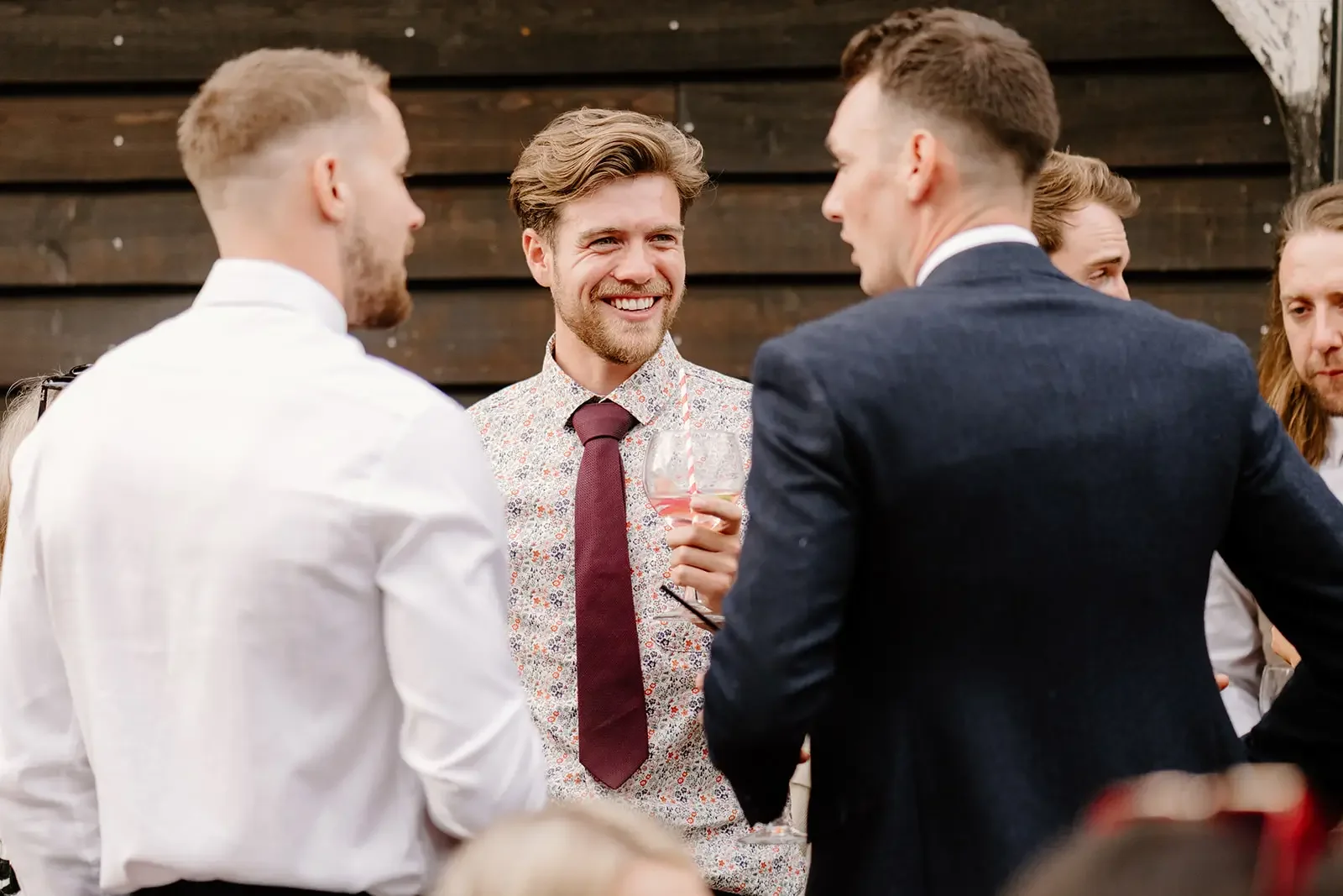 A group of people at a social gathering, engaging in conversation. The focus is on a man smiling, holding a glass of wine, while others around him are participating in the social event.