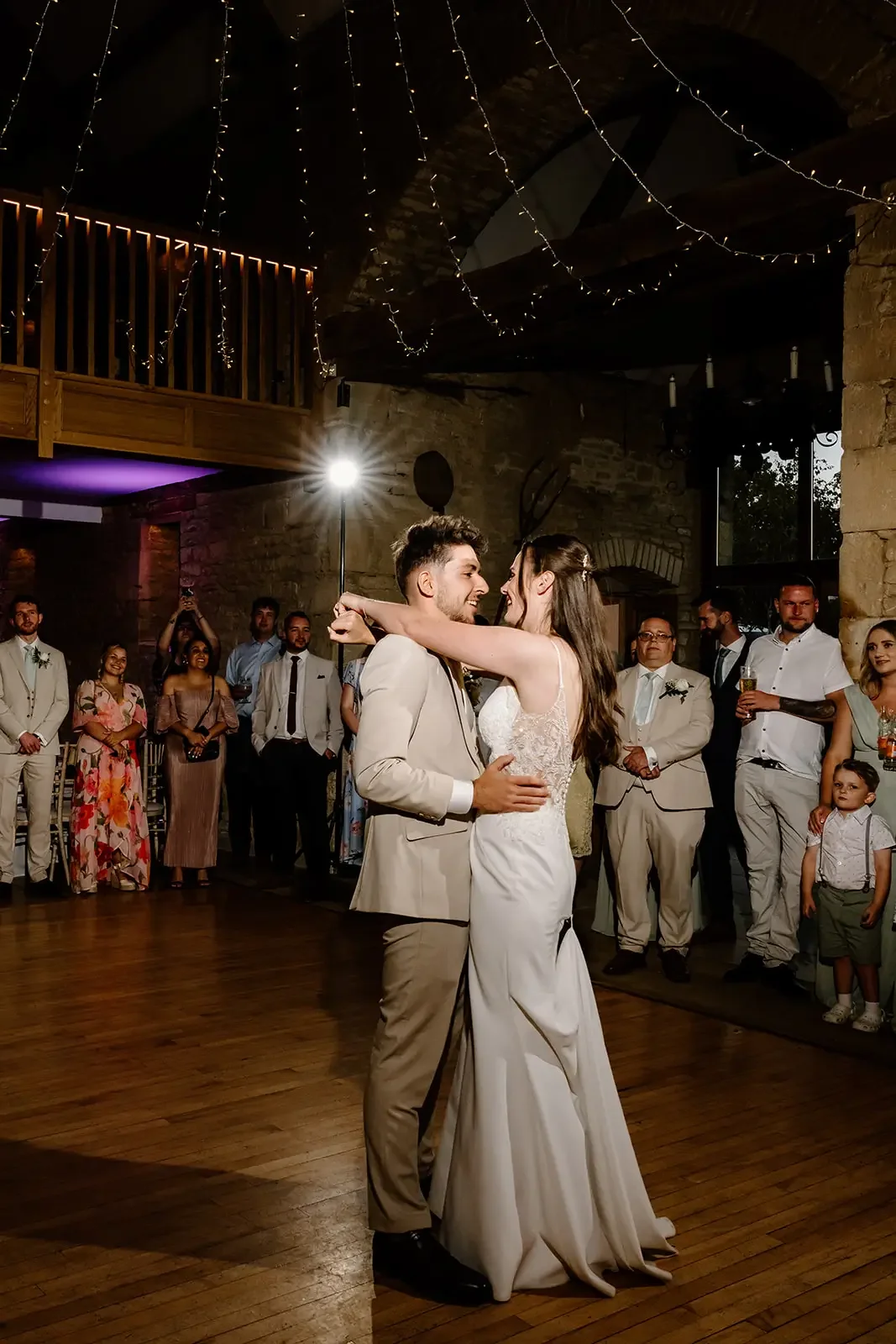A bride and groom dancing at their wedding reception, surrounded by friends and family, in a rustic venue decorated with fairy lights.
