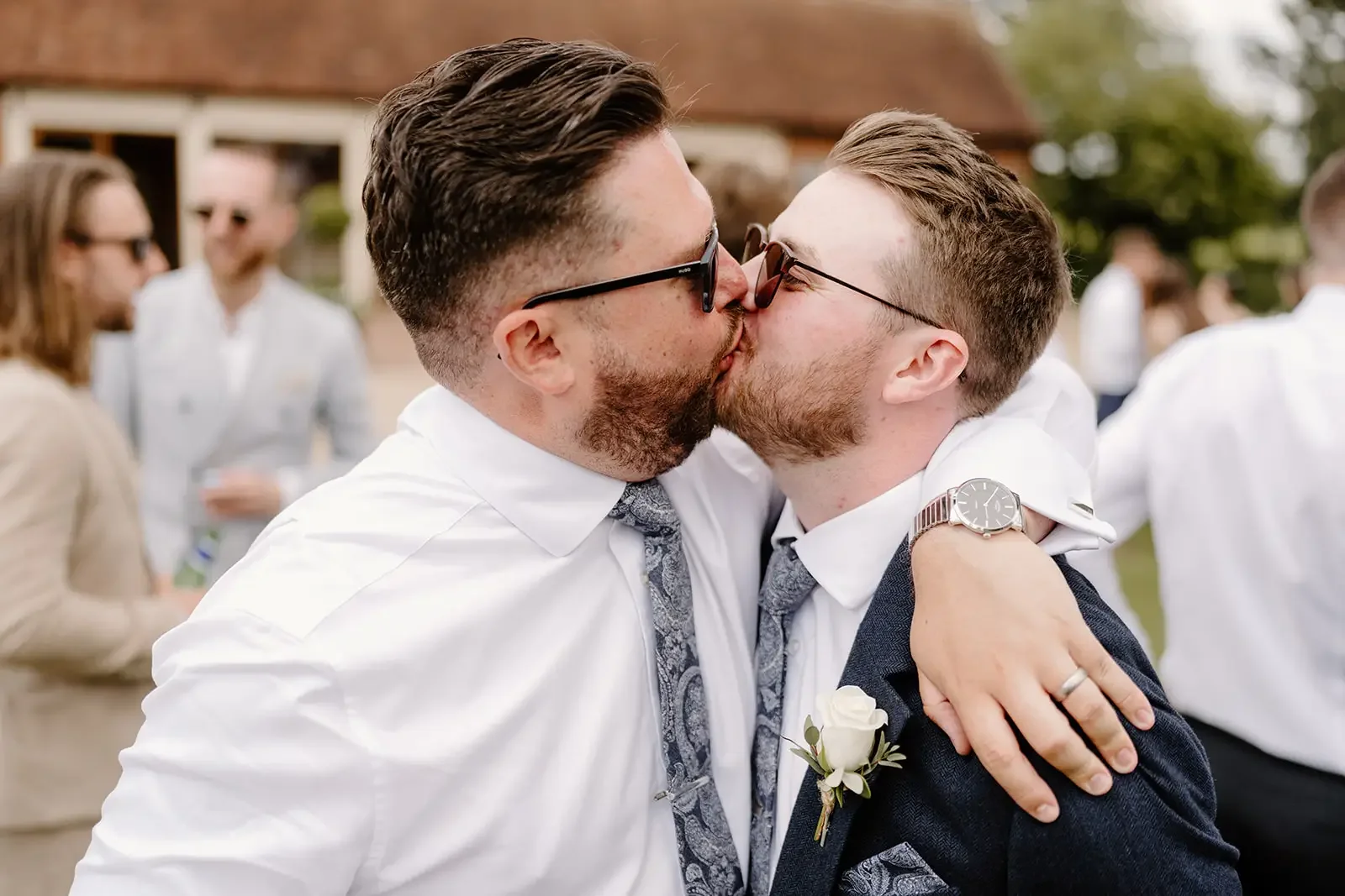 Two men wearing business suits and glasses sharing a kiss at an outdoor event, with a woman and other people in the background.