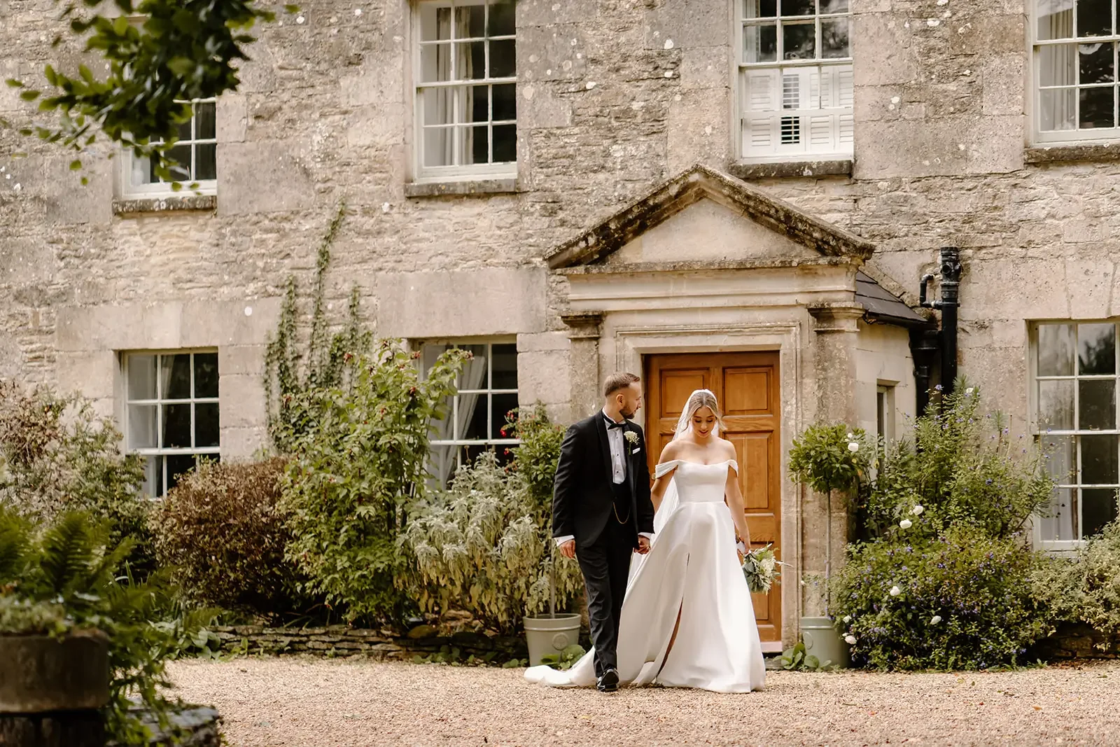 Bride and groom walking hand in hand outside a stone building with large windows and a wooden door, surrounded by greenery.