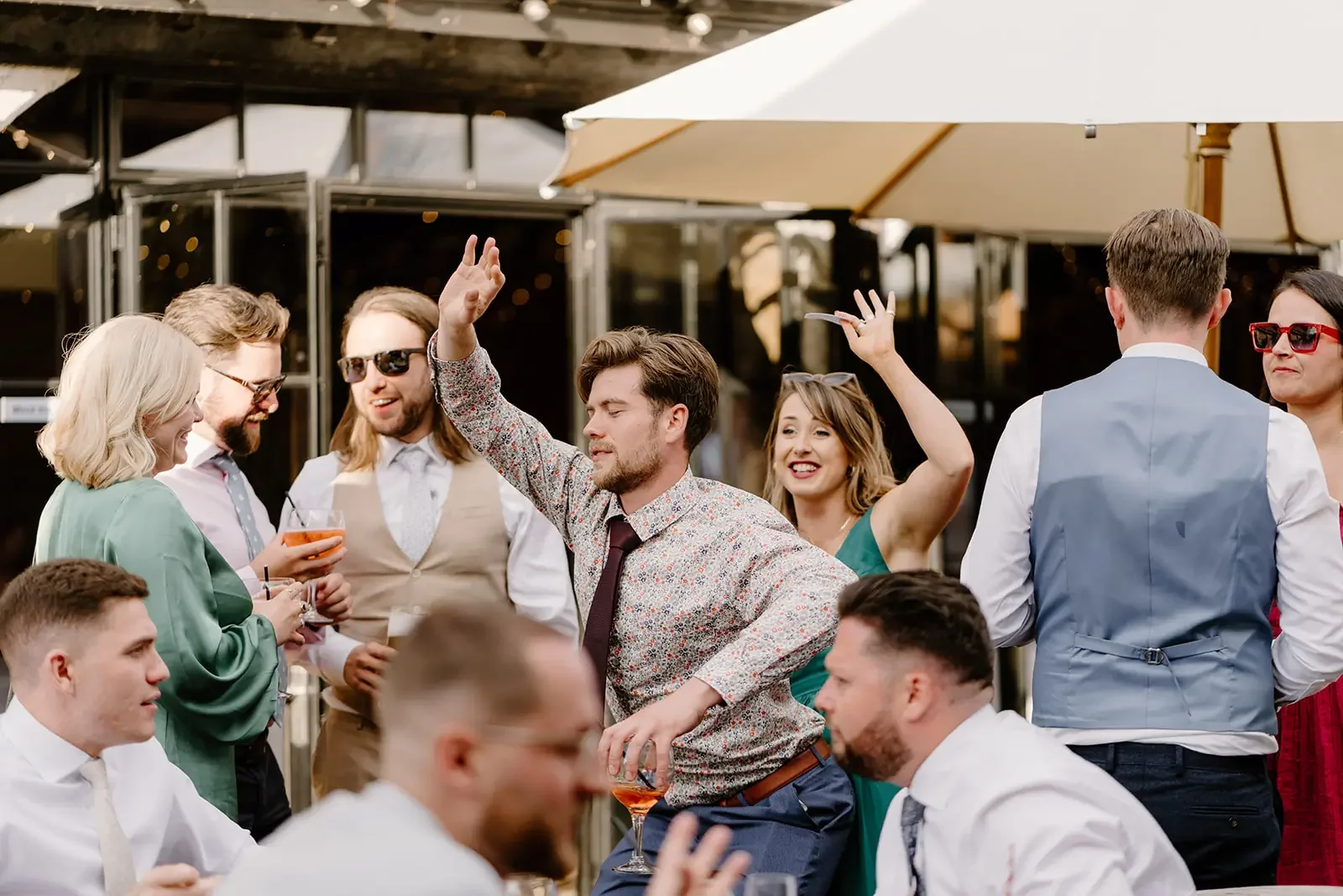 A group of people socializing at an outdoor event, some dancing and others talking, with drinks in hand, under a large umbrella.