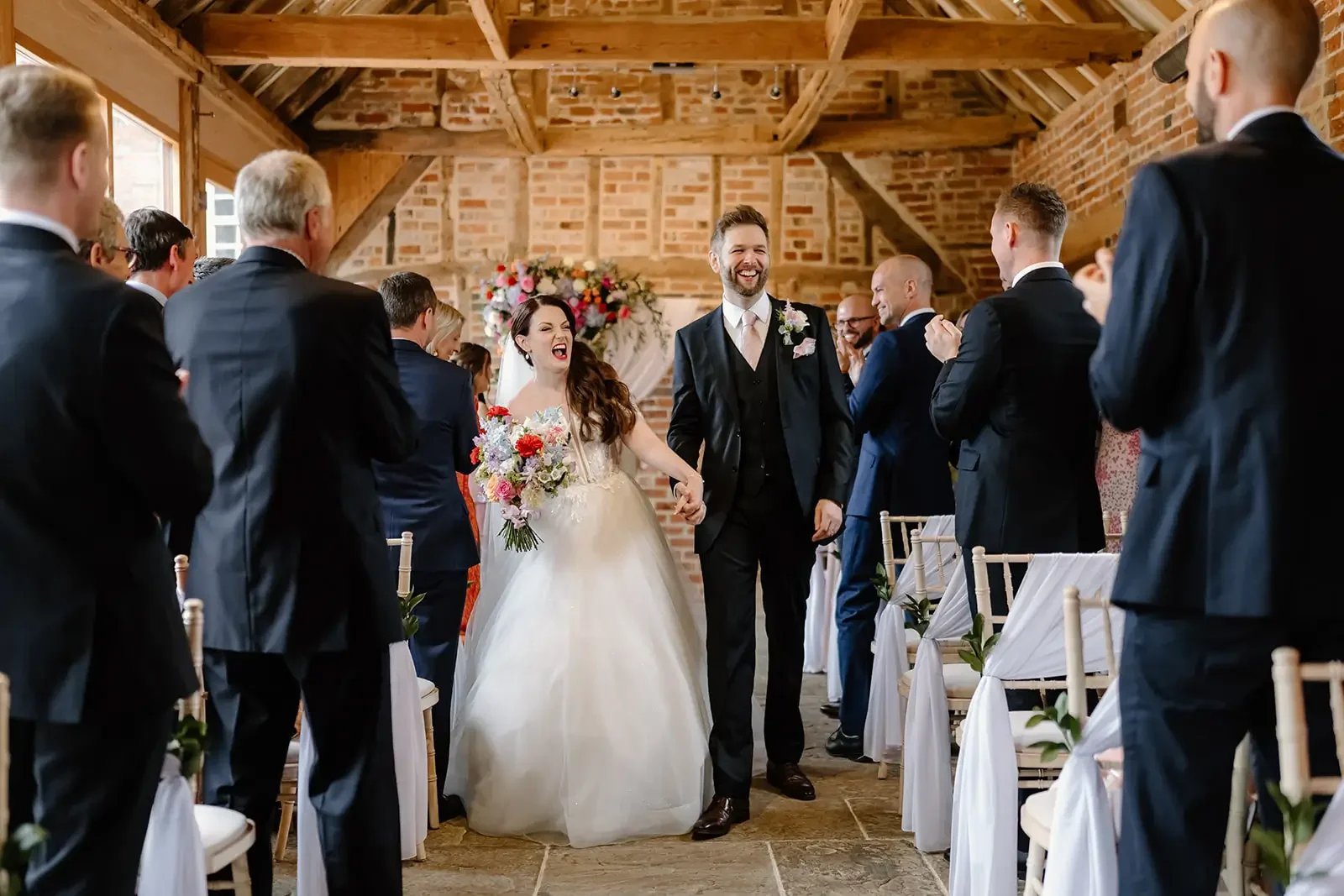A newly married couple walking down the aisle in a rustic wedding venue with exposed brick and wooden beams, surrounded by smiling wedding guests.