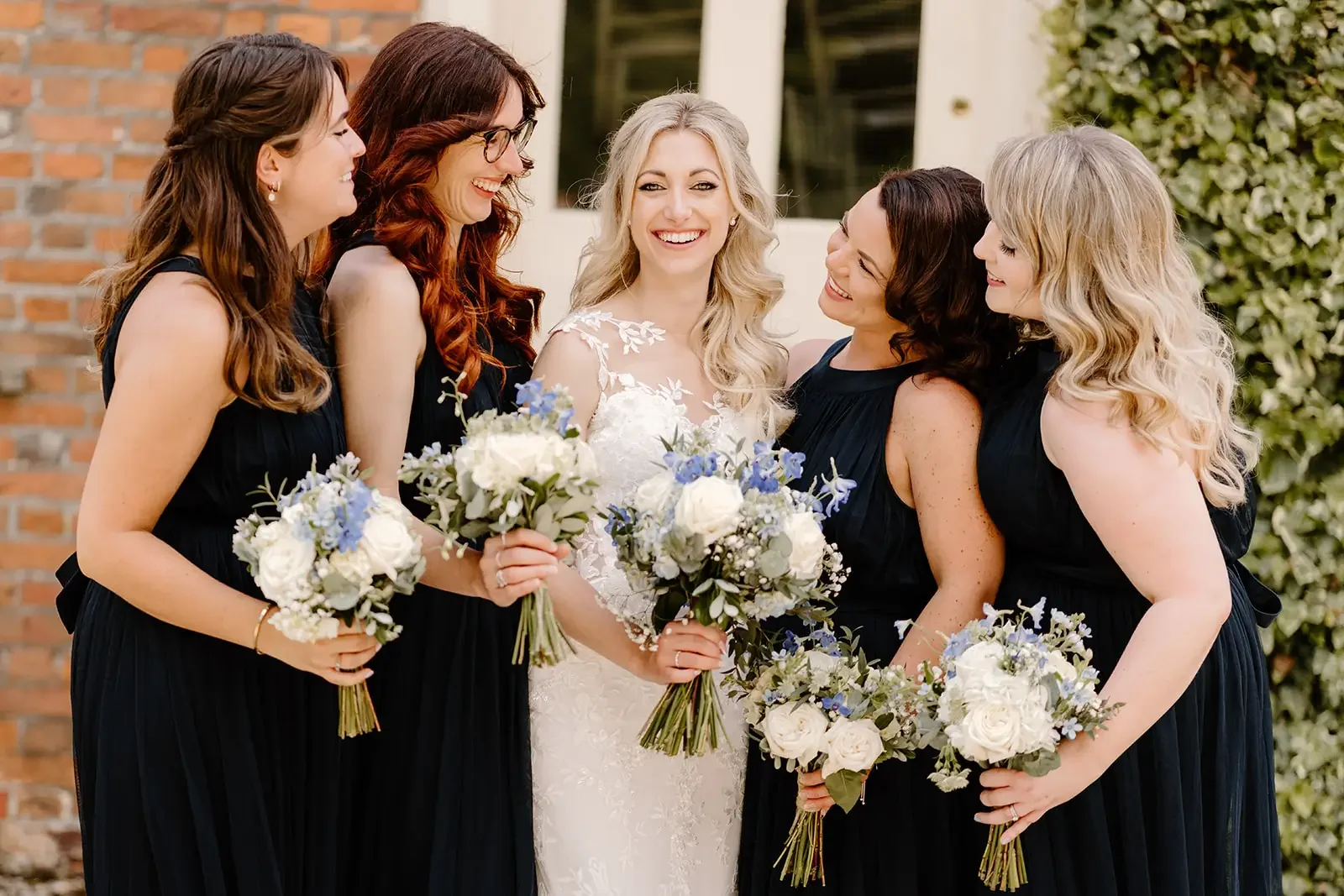 A bride in a white wedding dress holding a bouquet, surrounded by five women in black dresses holding bouquets, smiling and talking outdoors.
