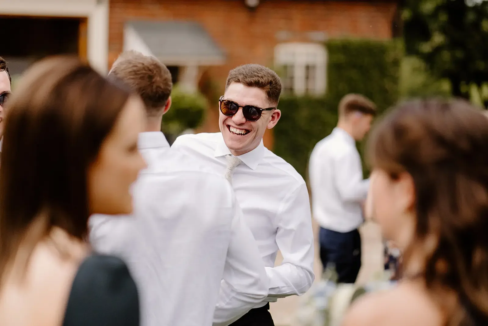 A group of people at an outdoor gathering, with a young man in sunglasses and a white dress shirt smiling and talking to others.